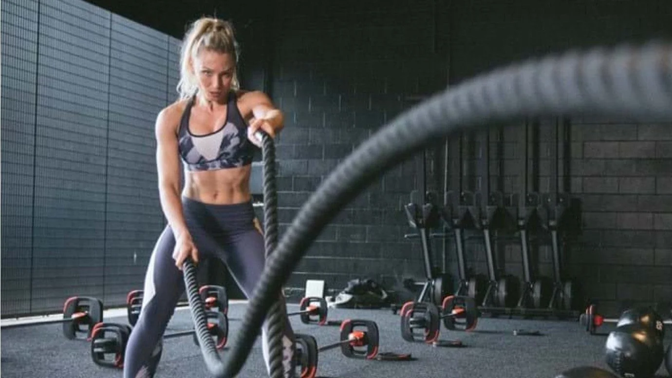 Woman exercising with battle ropes in a gym, with kettlebells and exercise bikes in the background.