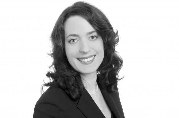 A professional black-and-white headshot of a woman with wavy dark hair, wearing a dark blazer and a small necklace, smiling at the camera.