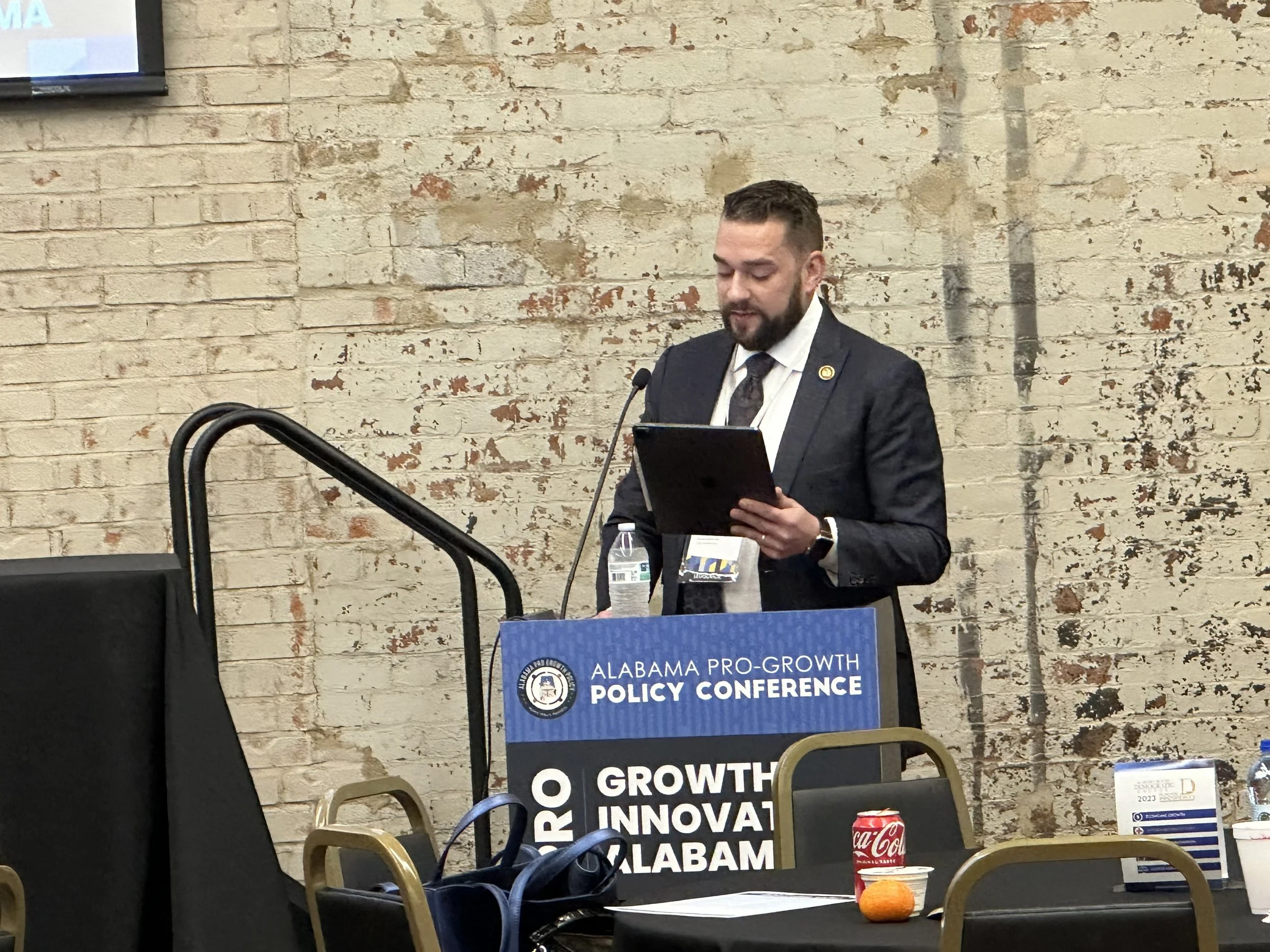A man with a beard and short hair standing at a podium, reading from a tablet during the Alabama Pro-Growth Policy Conference, with a brick wall in the background. The podium has a sign that reads 'Alabama Pro-Growth Policy Conference' and 'Growth Innovation Alabama.' There are water bottles, a Coca-Cola can, a small orange, and other conference materials on the table.