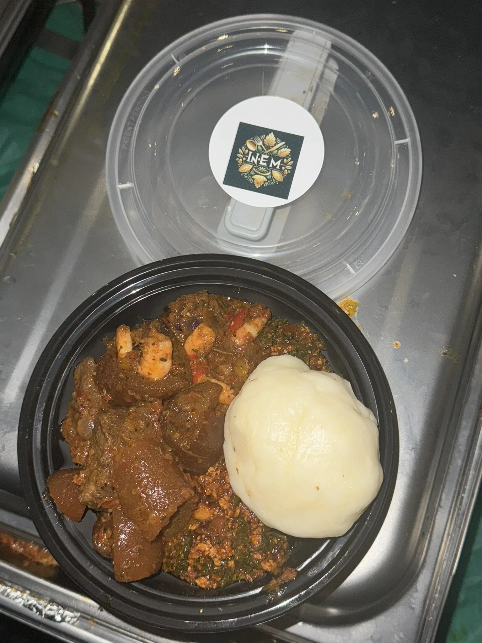 A black plastic container with beef stew, a serving of mashed potatoes, and vegetable mixture, placed on a metal surface with a clear plastic lid nearby.