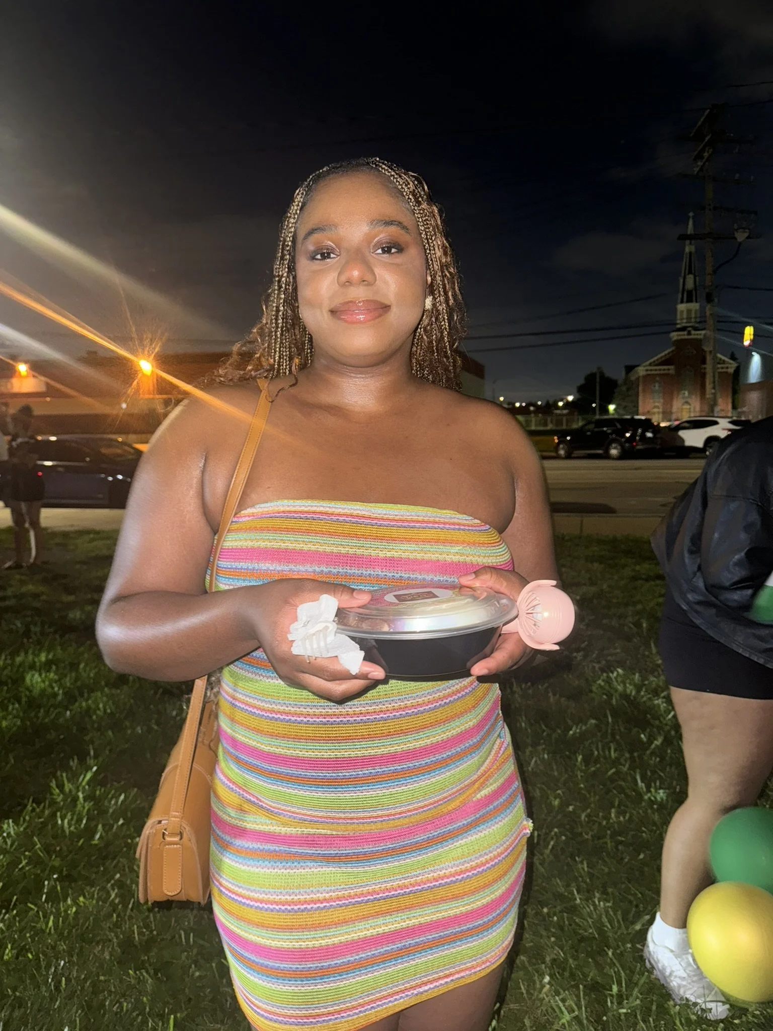 Woman wearing a colorful strapless dress holding a food container and a small pink balloon, standing outdoors at night with streetlights and cars in the background.