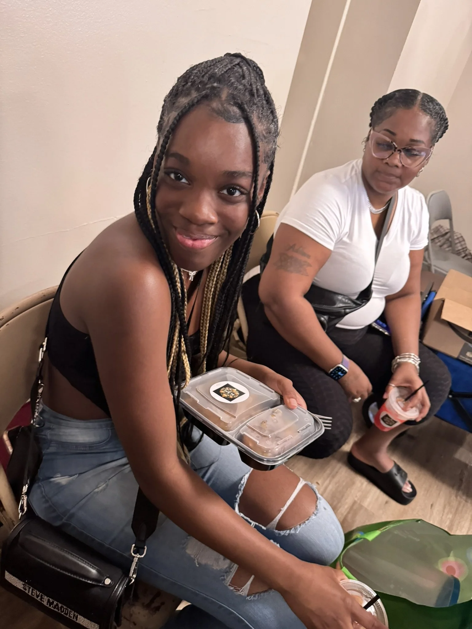Two women sitting at a table, with the younger woman holding a food container, and the older woman holding a drink. The younger woman has braided hair and is smiling at the camera. The older woman is wearing glasses and a white shirt, looking to the 