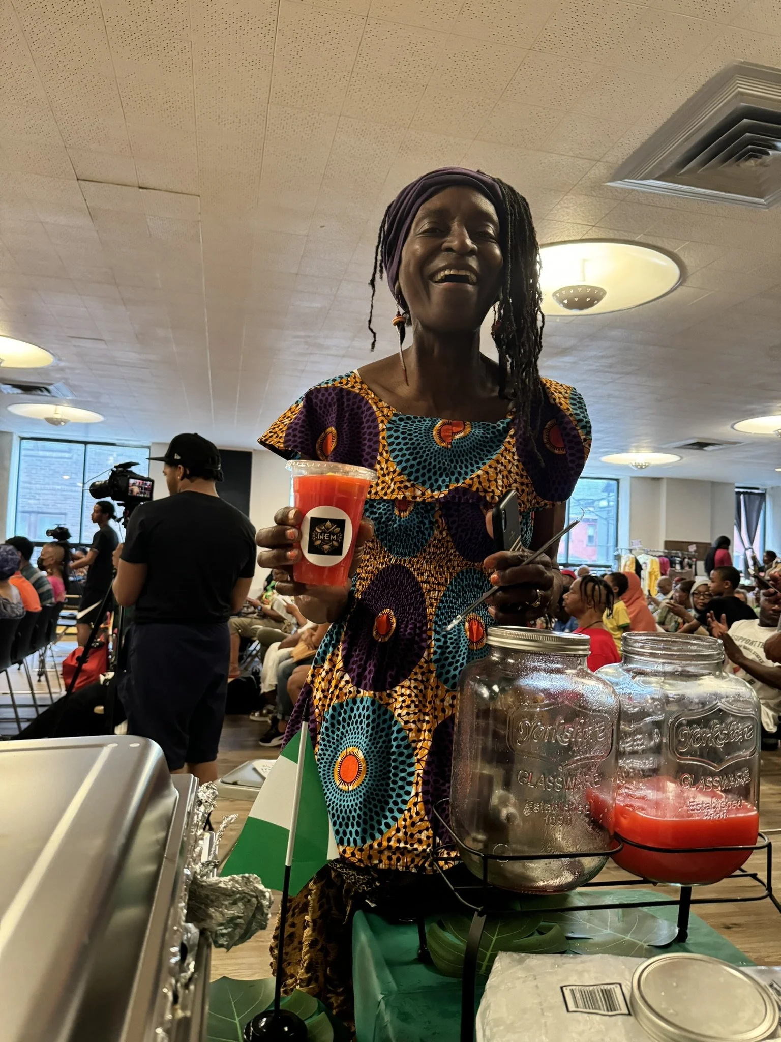 A woman with dreadlocks and a colorful African-patterned dress smiling and holding a red beverage in a clear cup at an indoor event. She is standing behind a table with jars of juice and an Italian flag, with many people seated and a camera crew in t