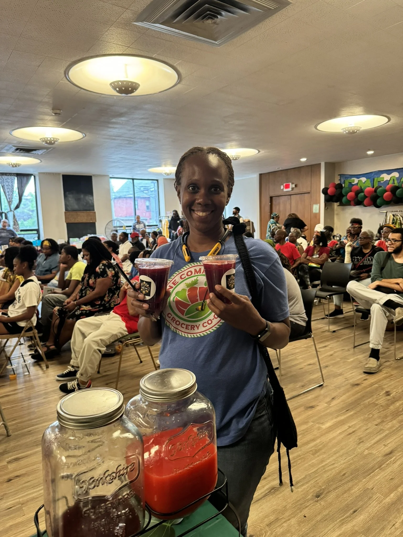 A woman smiling and holding two cups of purple juice at a community event. In the foreground, two large jars of red-colored juice are visible. The event has many attendees seated in chairs in a brightly lit room decorated with balloons and a banner.