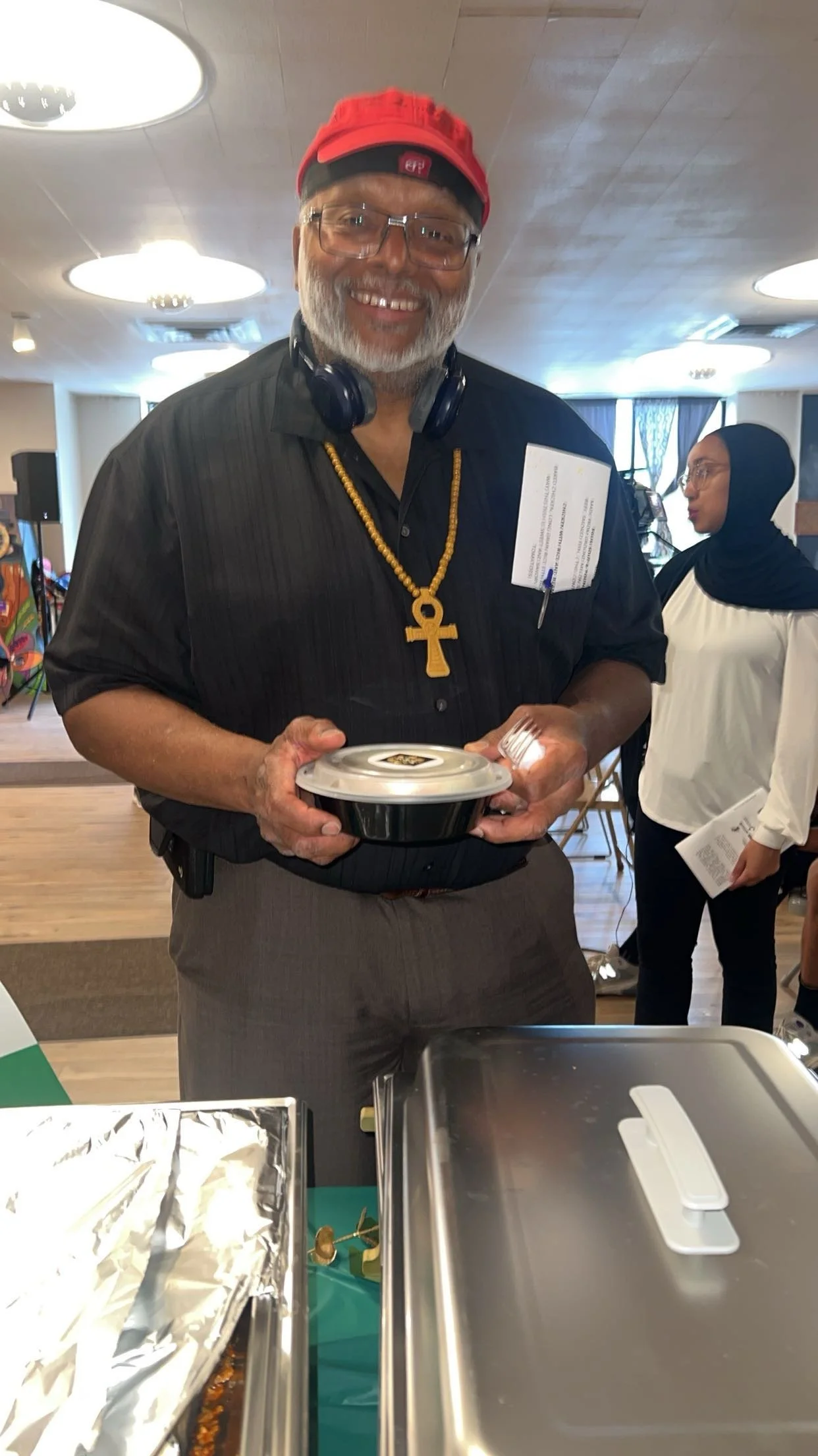 Man with gray beard wearing glasses, a red hat, and a large ankh necklace, holding a takeout container and smiling. In the background, a woman with a black headscarf and white blouse, holding a program or pamphlet, standing in a room with tables and 