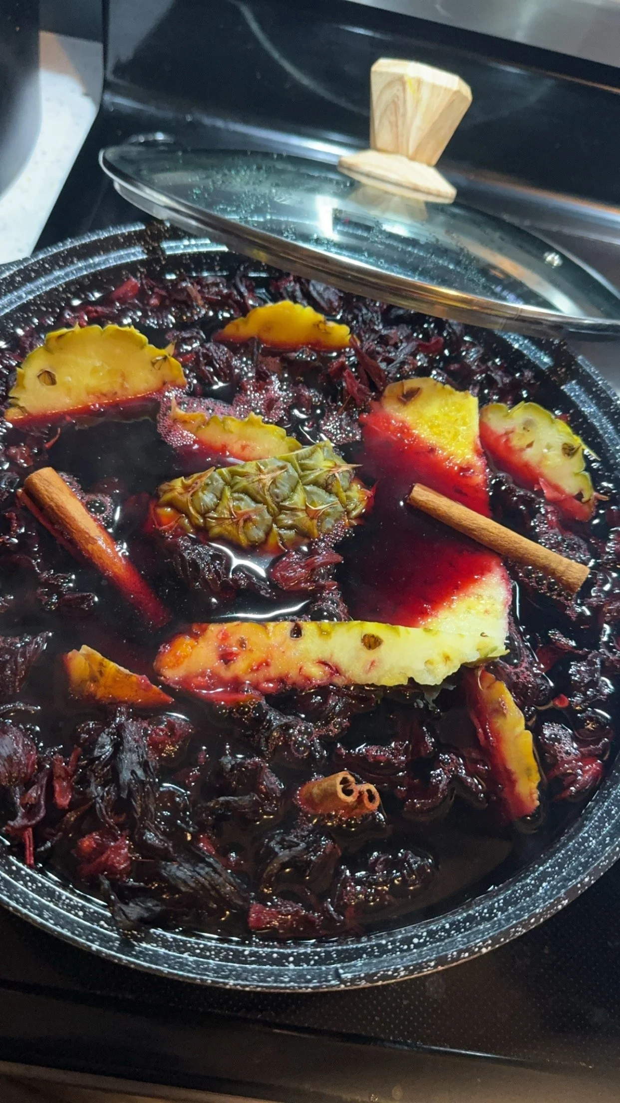 A pot on a stovetop filled with a dark liquid, pineapple slices, cinnamon sticks, and other spices, possibly making mulled wine or similar beverage.