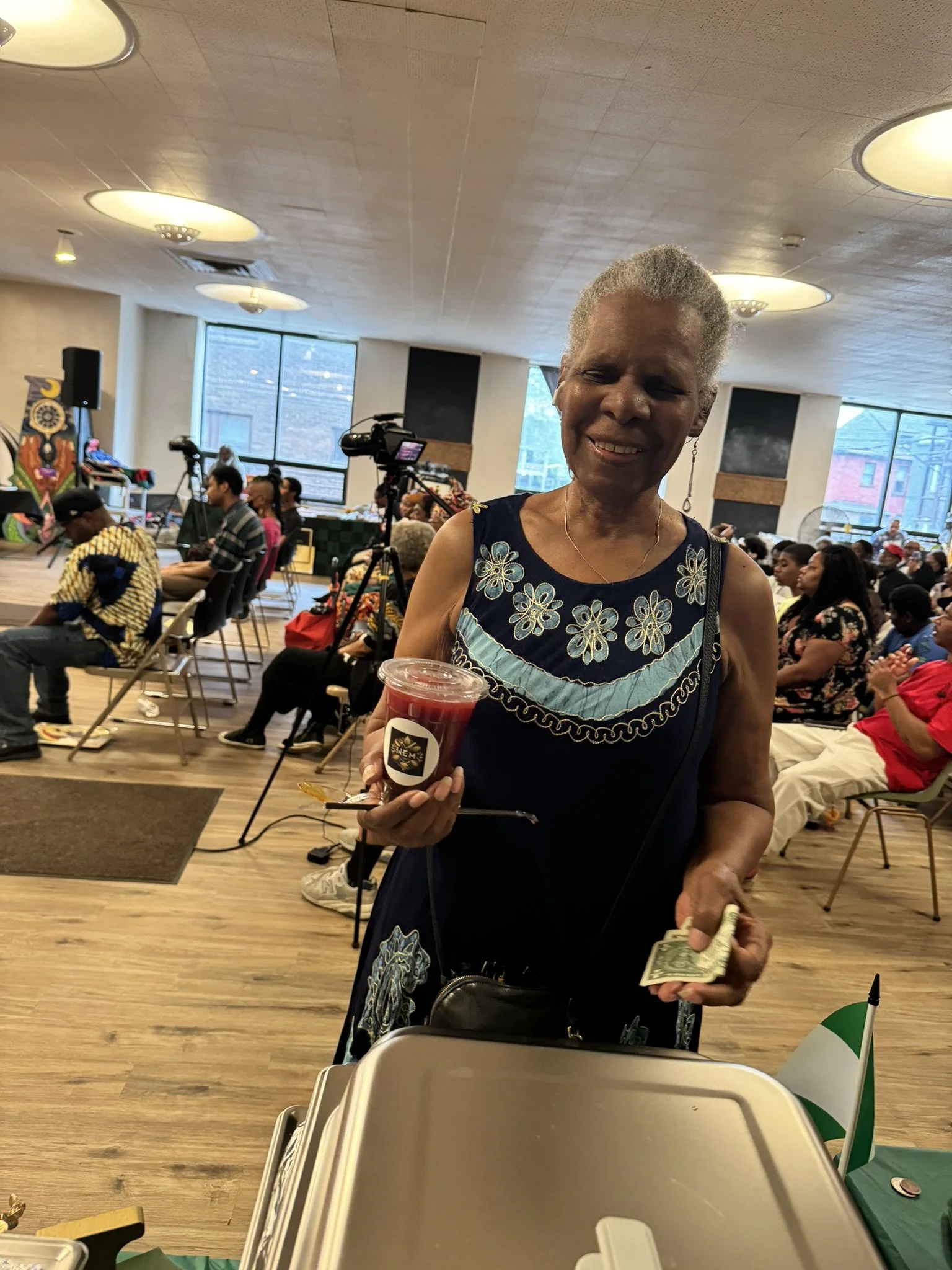 A smiling elderly woman holding a cup of red beverage and cash in her hands at an indoor event with many seated attendees and a stage.