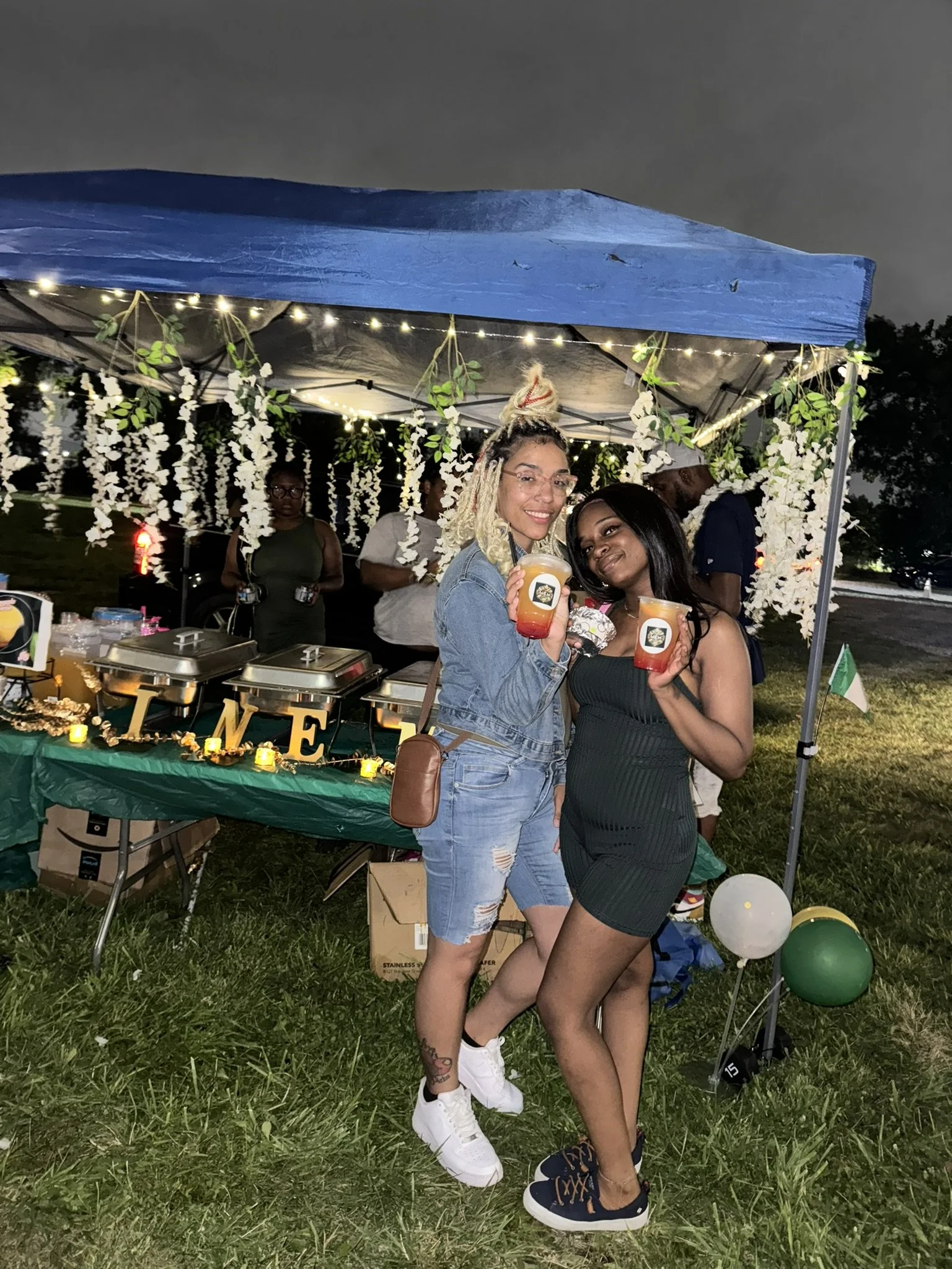 Two women posing with drinks at an outdoor event or party under a decorated canopy, with a table of food and decorations behind them, illuminated by string lights.
