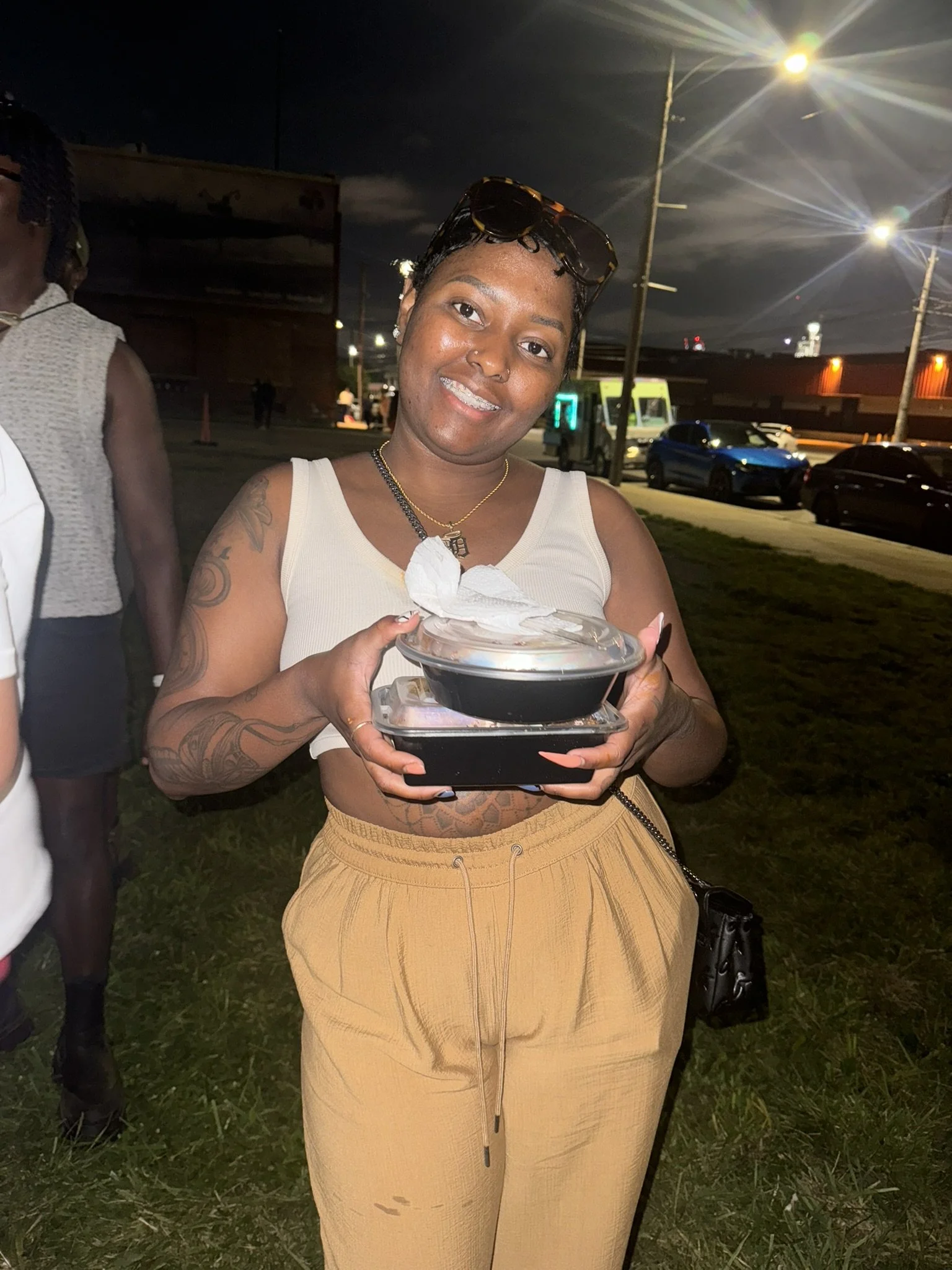 A woman standing outdoors at night, holding a takeout food container, smiling at the camera, wearing a sleeveless beige top and yellow pants.