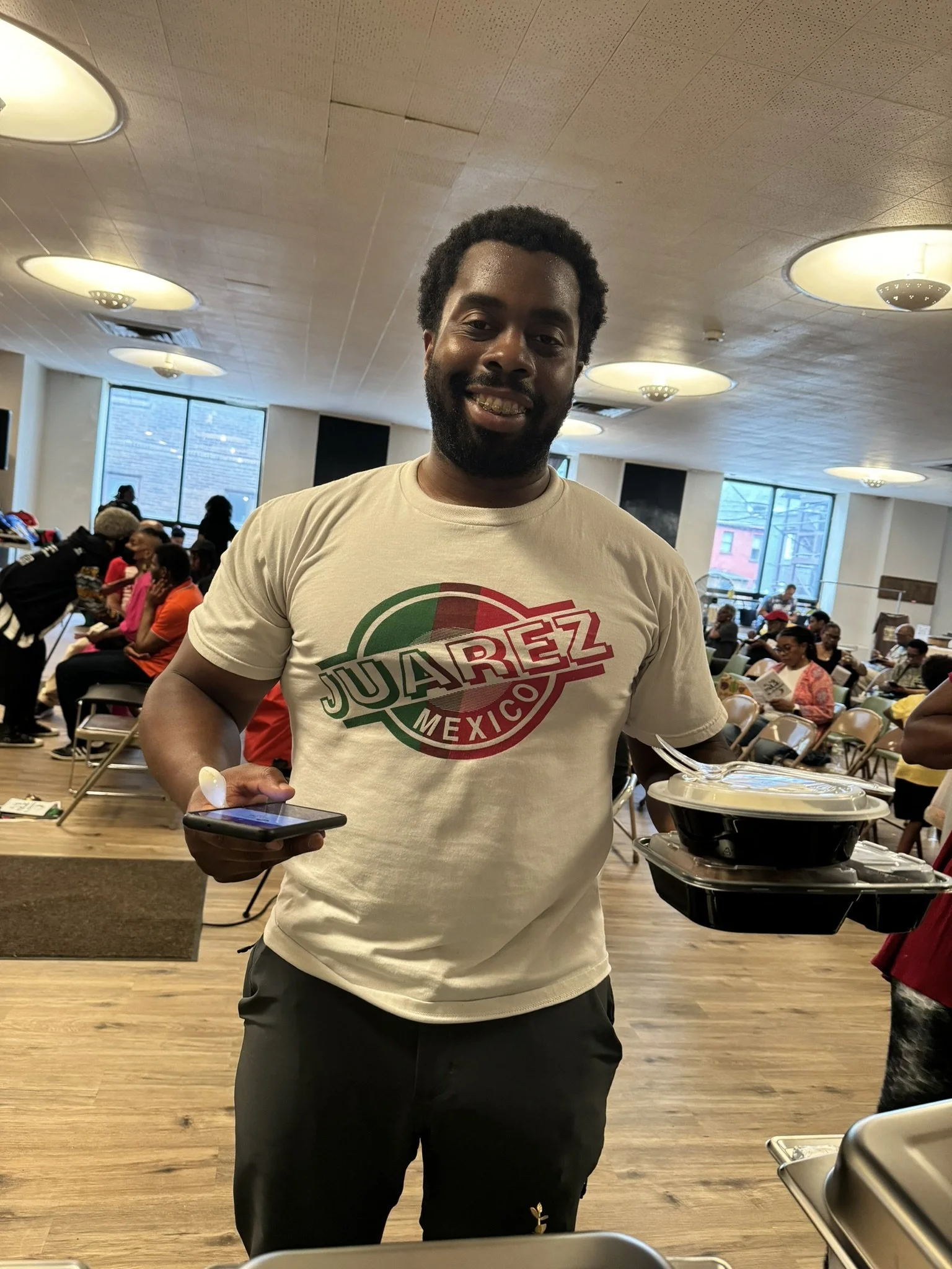 A man with dark hair and beard smiling, holding a smartphone in one hand and a black takeout container in the other, standing in a busy dining hall with other people seated at tables.