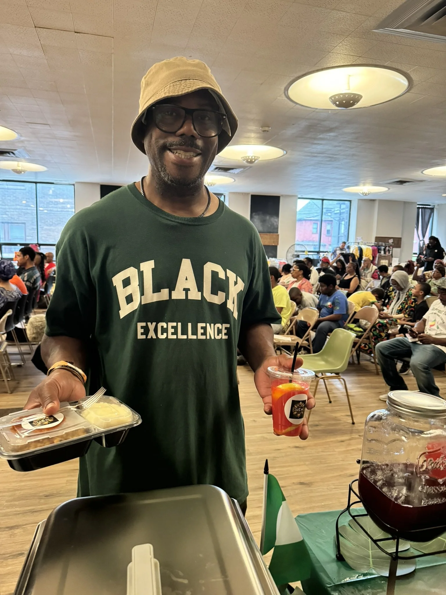 A man wearing a tan bucket hat, glasses, and a green shirt with 'BLACK EXCELLENCE' printed on it standing at a community event, holding a packaged meal in one hand and a cup of red beverage with fruit slices and a straw in the other. There are many p