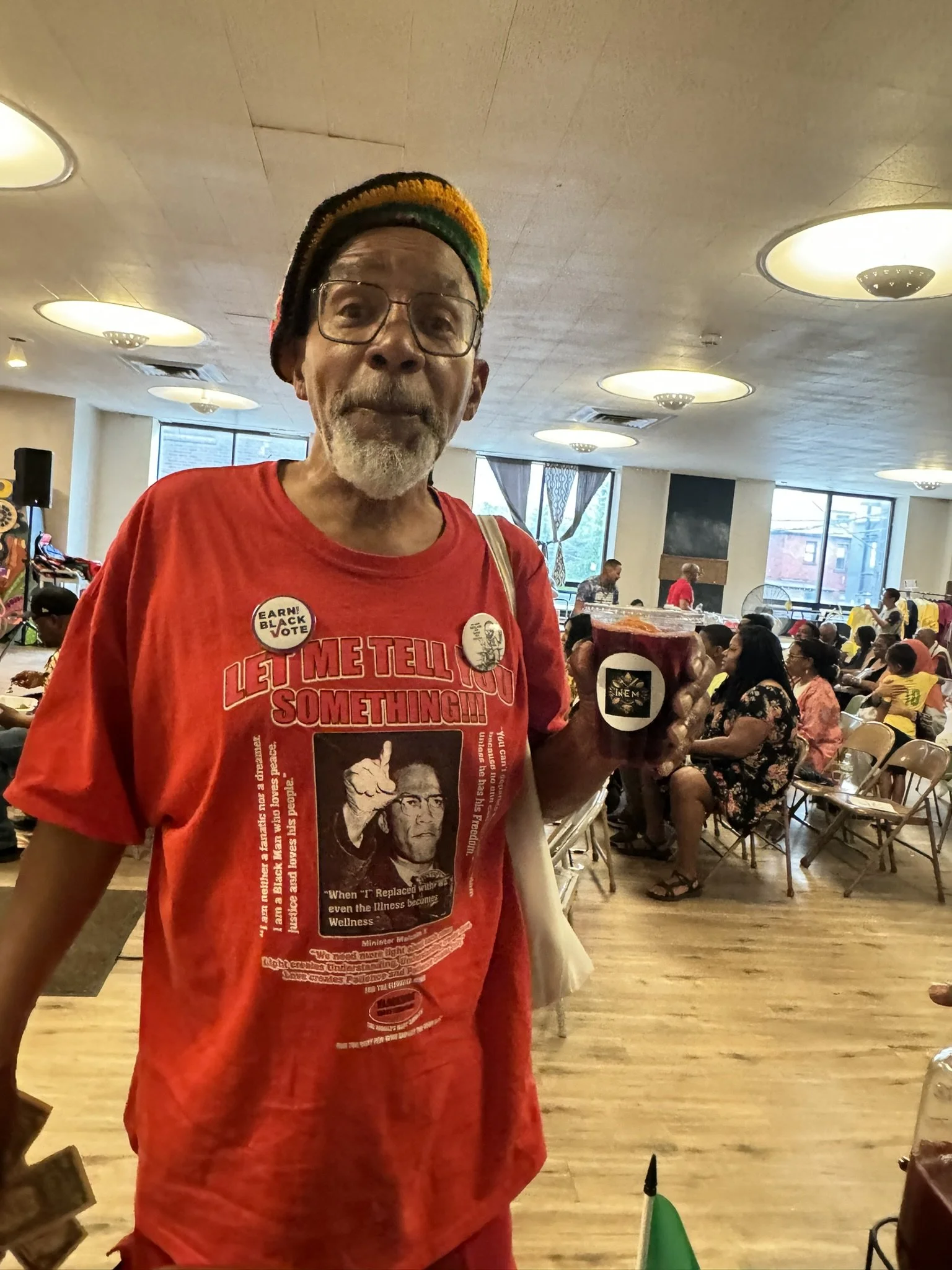 An older man with glasses, a gray beard, and wearing a rainbow-colored beanie and red T-shirt, holding a cup of red beverage, standing in a crowded indoor space with people sitting on chairs and tables.