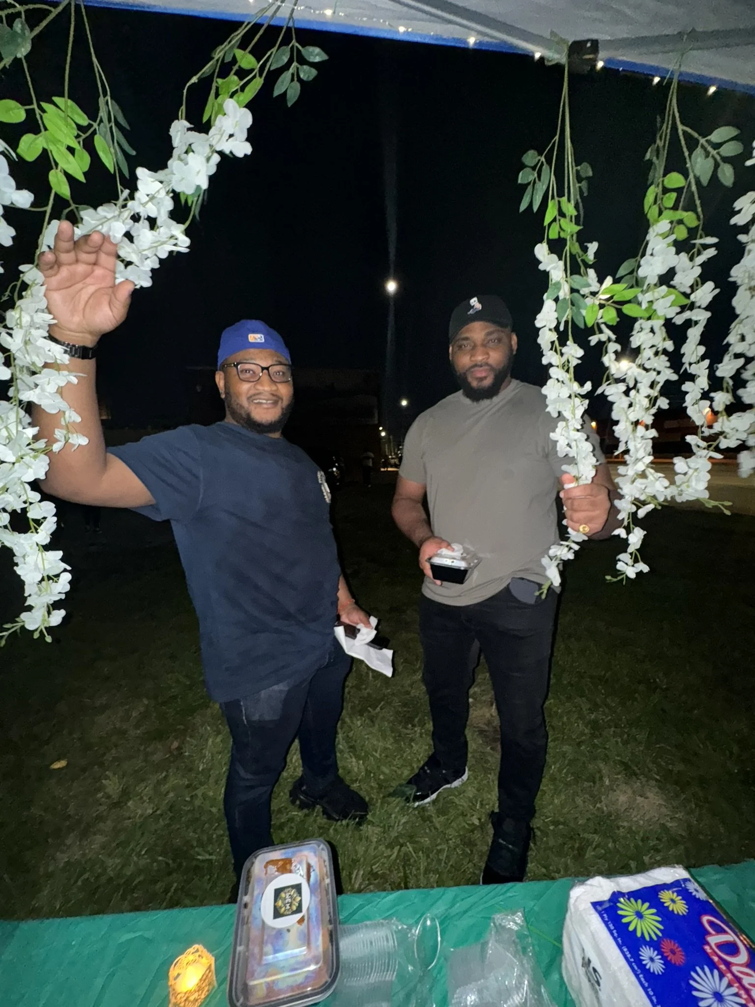 Two men standing outdoors at night, framed by hanging white flowers, smiling at the camera. One man wears a blue shirt and cap, the other a gray shirt and black cap. There is a table in front with a container of food and a pack of tissues.