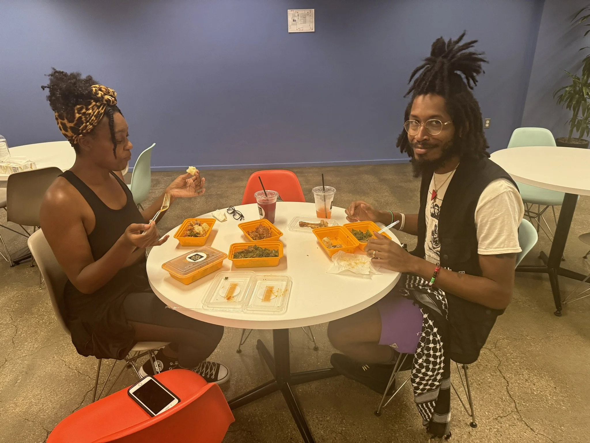 A woman and a man sitting at a round table eating takeout food in a room with a blue wall. The woman wears a black dress and leopard print headwrap, and the man wears glasses, a white t-shirt, and patterned pants. There are two cups with drinks, seve
