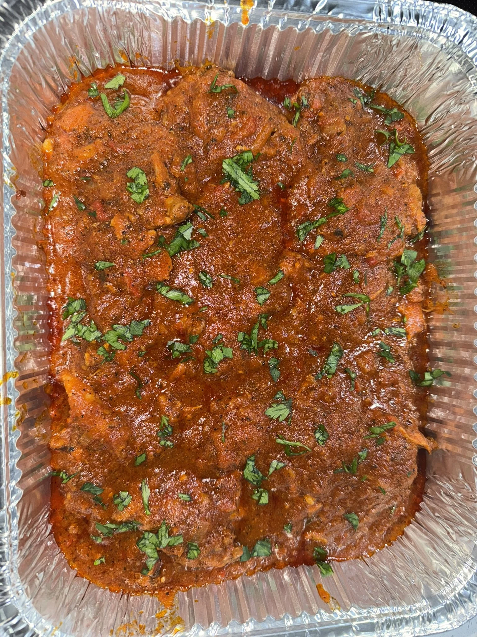 A foil container with cooked meat in a red sauce, garnished with chopped parsley.