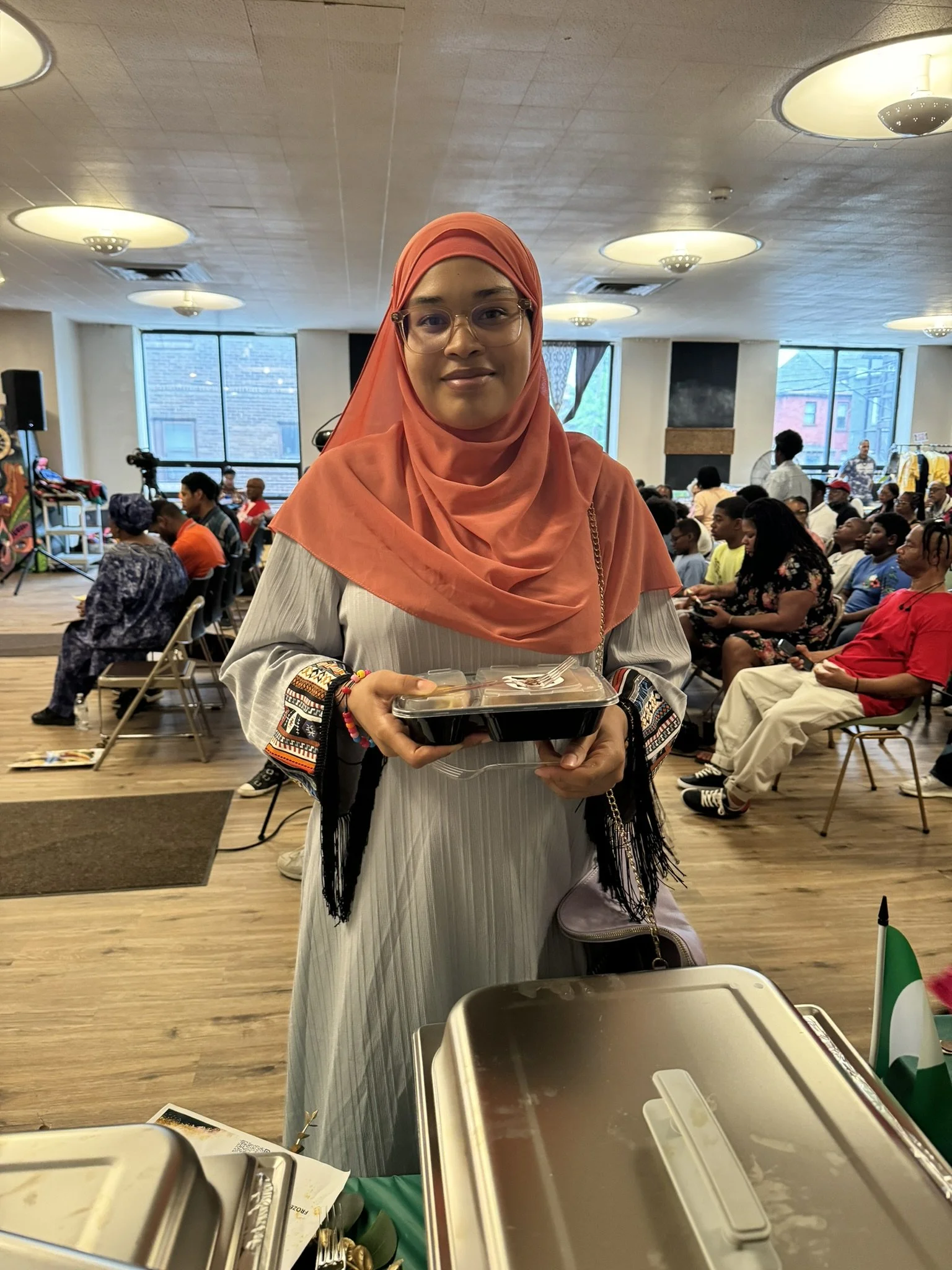 A woman wearing glasses and a coral hijab stands in a large hall, holding a food container and smiling at the camera. Behind her, there are several people seated and an event is taking place.