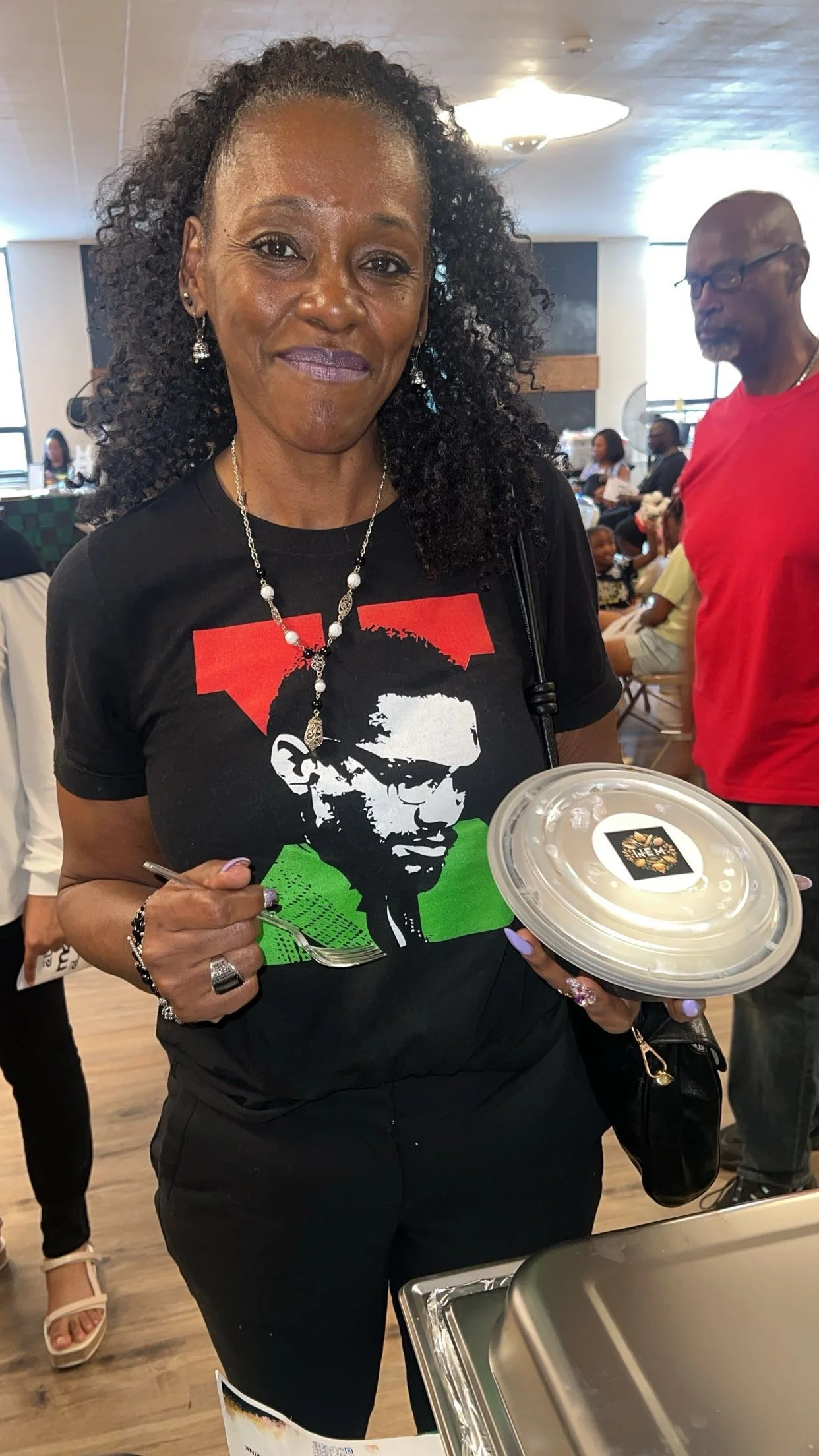 A woman with curly hair wearing a black t-shirt with a graphic of a man with sunglasses in red, black, and green, holding a fork and a white plate with a logo, at a gathering or event. She is smiling and has accessories including a necklace, earrings