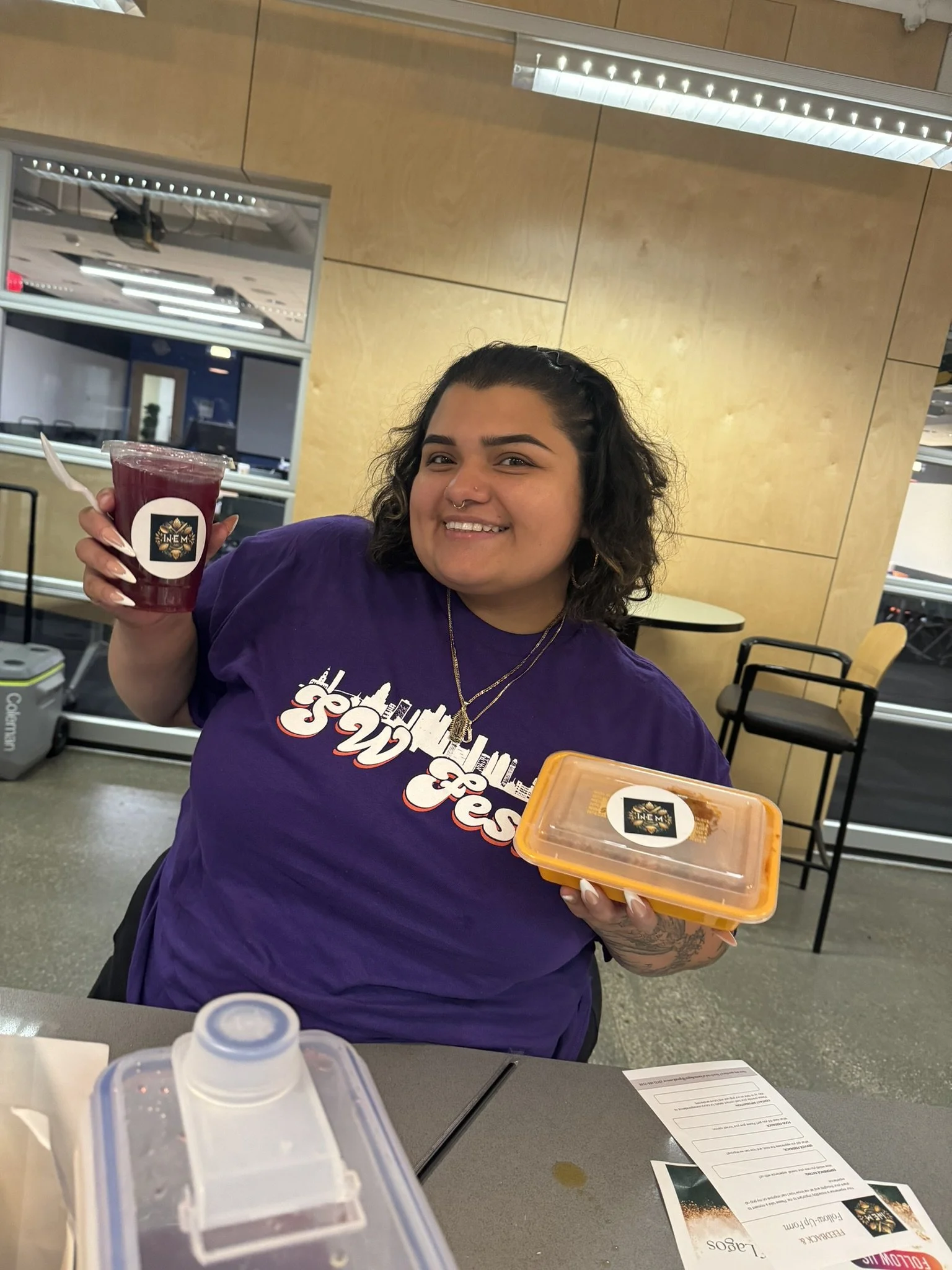 A woman with dark curly hair and a nose ring smiling at the camera, holding a purple beverage in a clear plastic cup with a sticker, and a yellow food container with a sticker. She is wearing a purple T-shirt with a city skyline graphic and text, in 