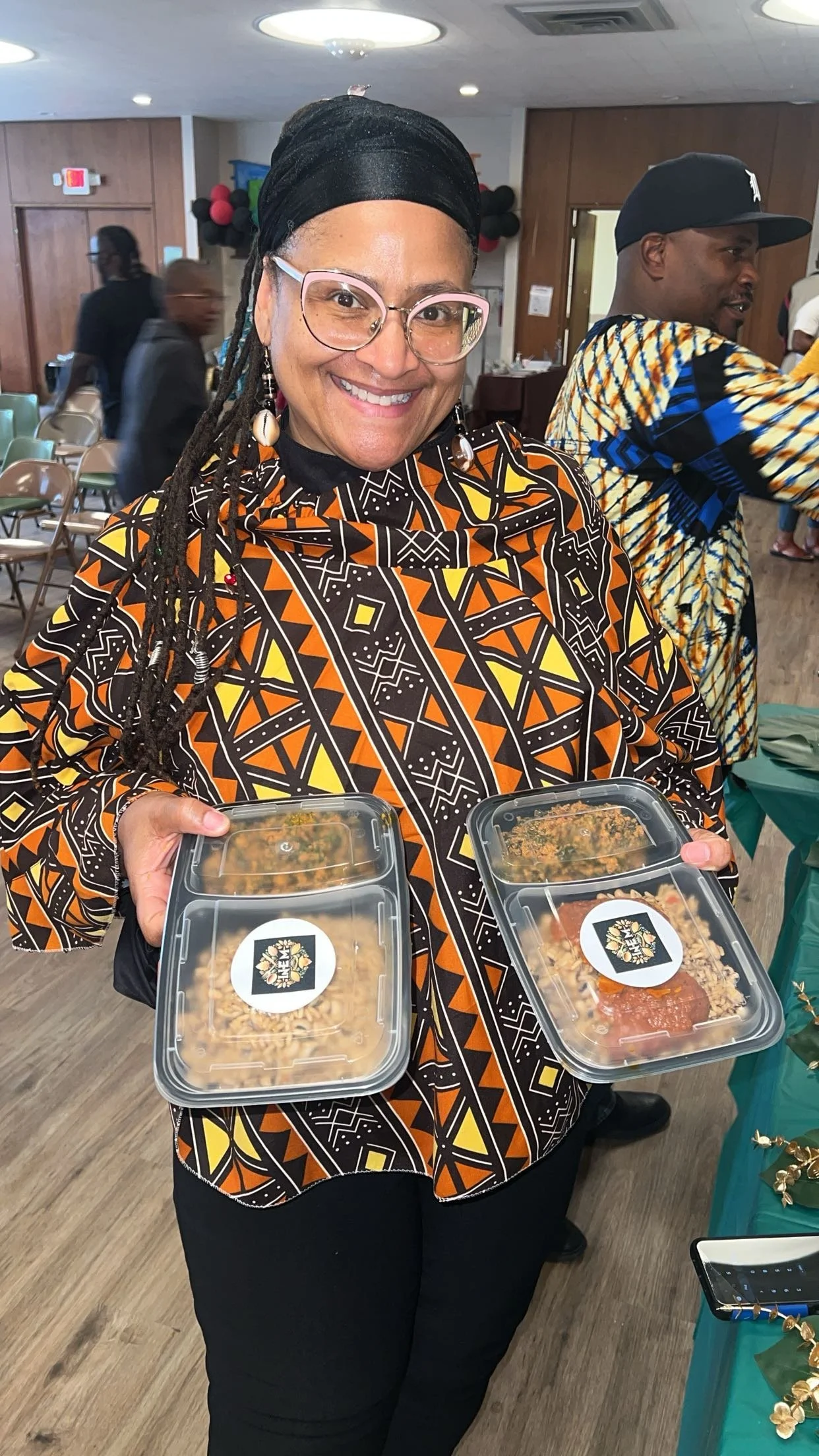 Smiling woman with glasses and hair in dreadlocks holding two plastic food containers, at indoor event with seated and standing people in the background.