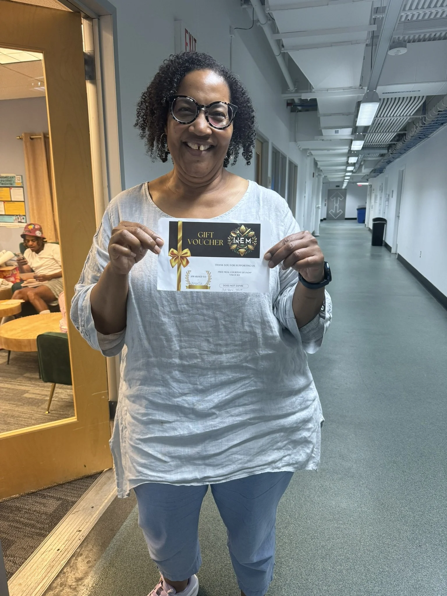 A woman with curly hair and glasses smiling while holding a gift voucher inside a building, with a hallway extending behind her.