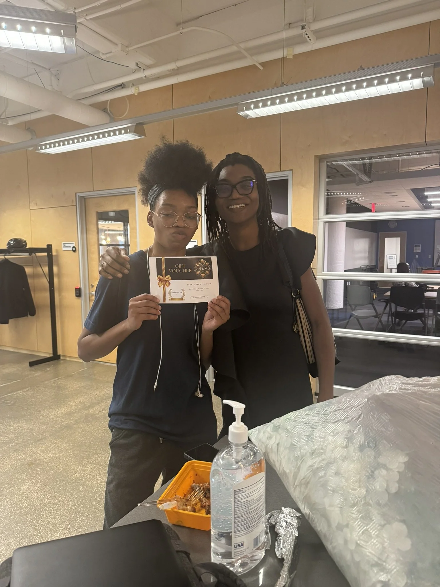 Two women stand together indoors, one holding a gift voucher with a bow design. They are smiling, and there are tables and chairs in the background.