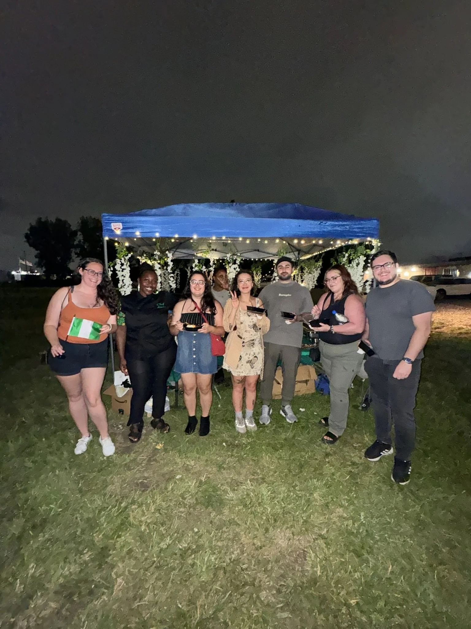 A group of nine people standing outdoors at night, posing for a photo under a decorated blue canopy with string lights, holding bowls and utensils, smiling, with grass ground and a dark, cloudy sky in the background.