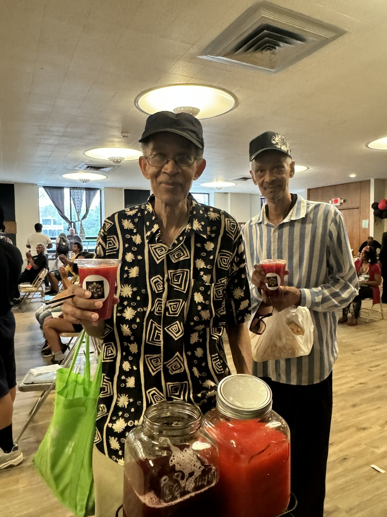 Two elderly men standing indoors, each holding a cup of red beverage, with jars of red and dark drink on the table in front of them. The man on the left is wearing a black cap and a patterned shirt, while the man on the right has a black hat and a st