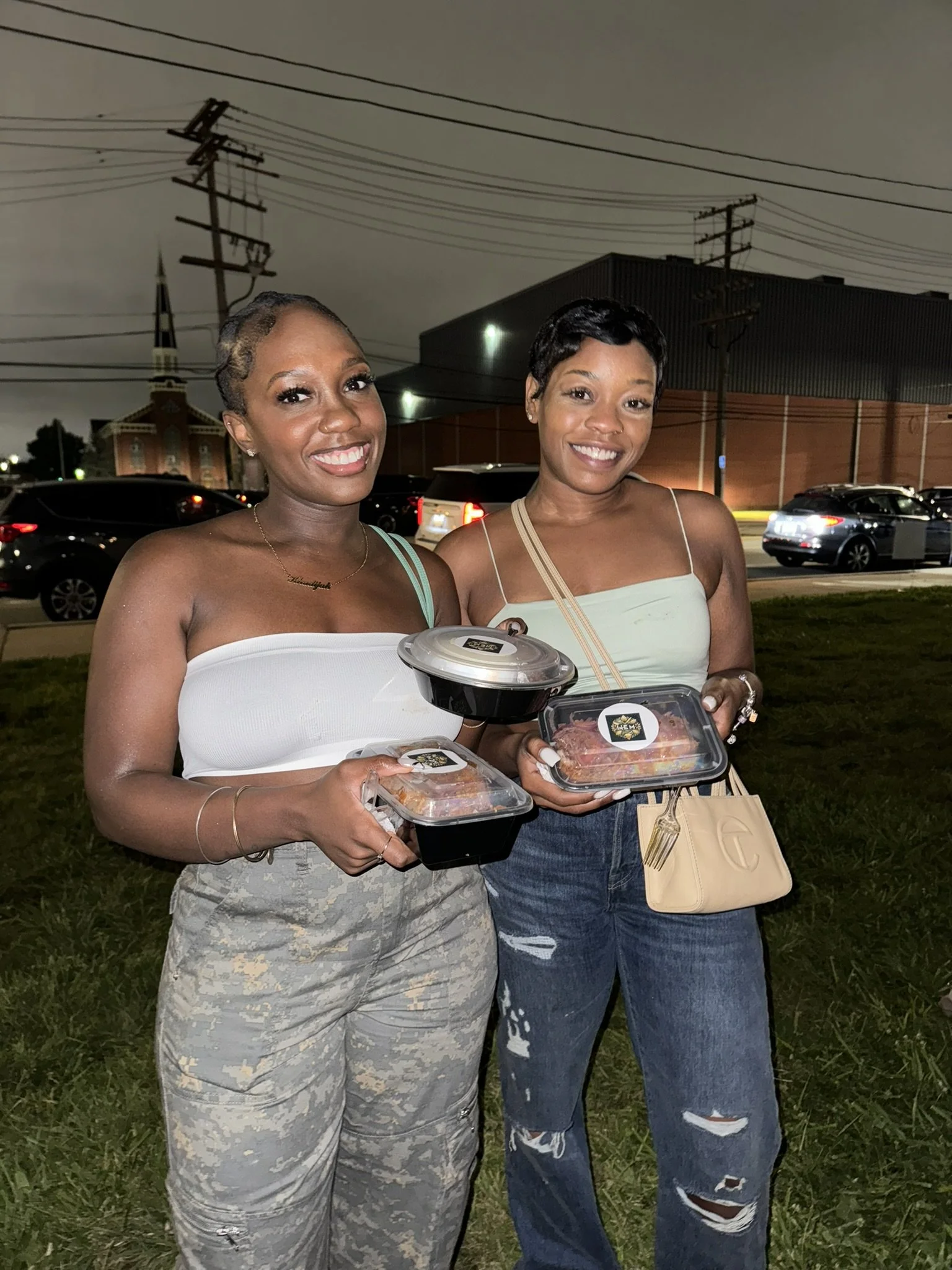 Two women standing outdoors at night, smiling, holding takeout containers of food, with cars and a building in the background.