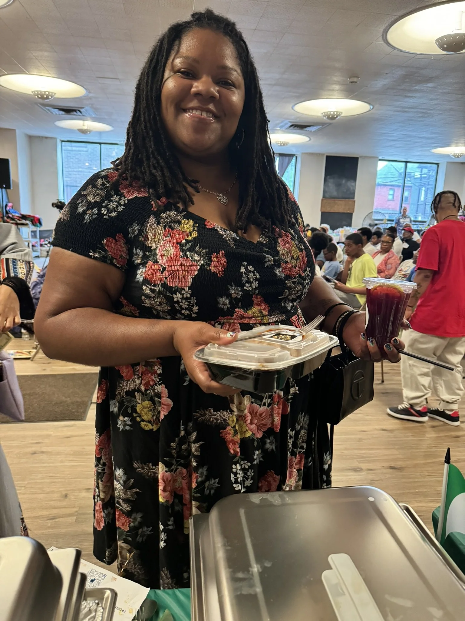 Smiling woman holding a takeout container and a cup of berries at an indoor event with many people in the background.