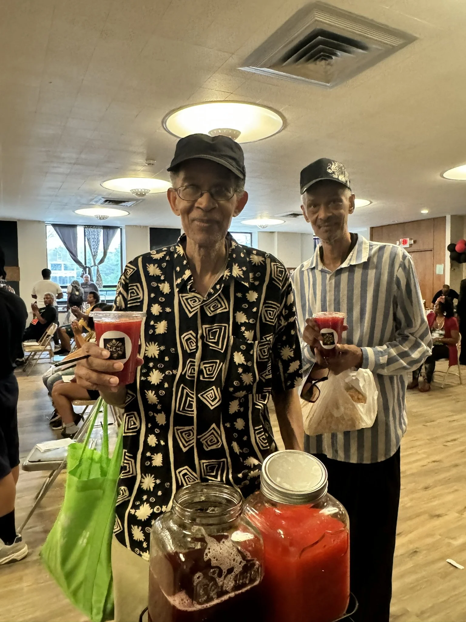 Two elderly men holding cups of red drinks inside a community center with people sitting and standing in the background.