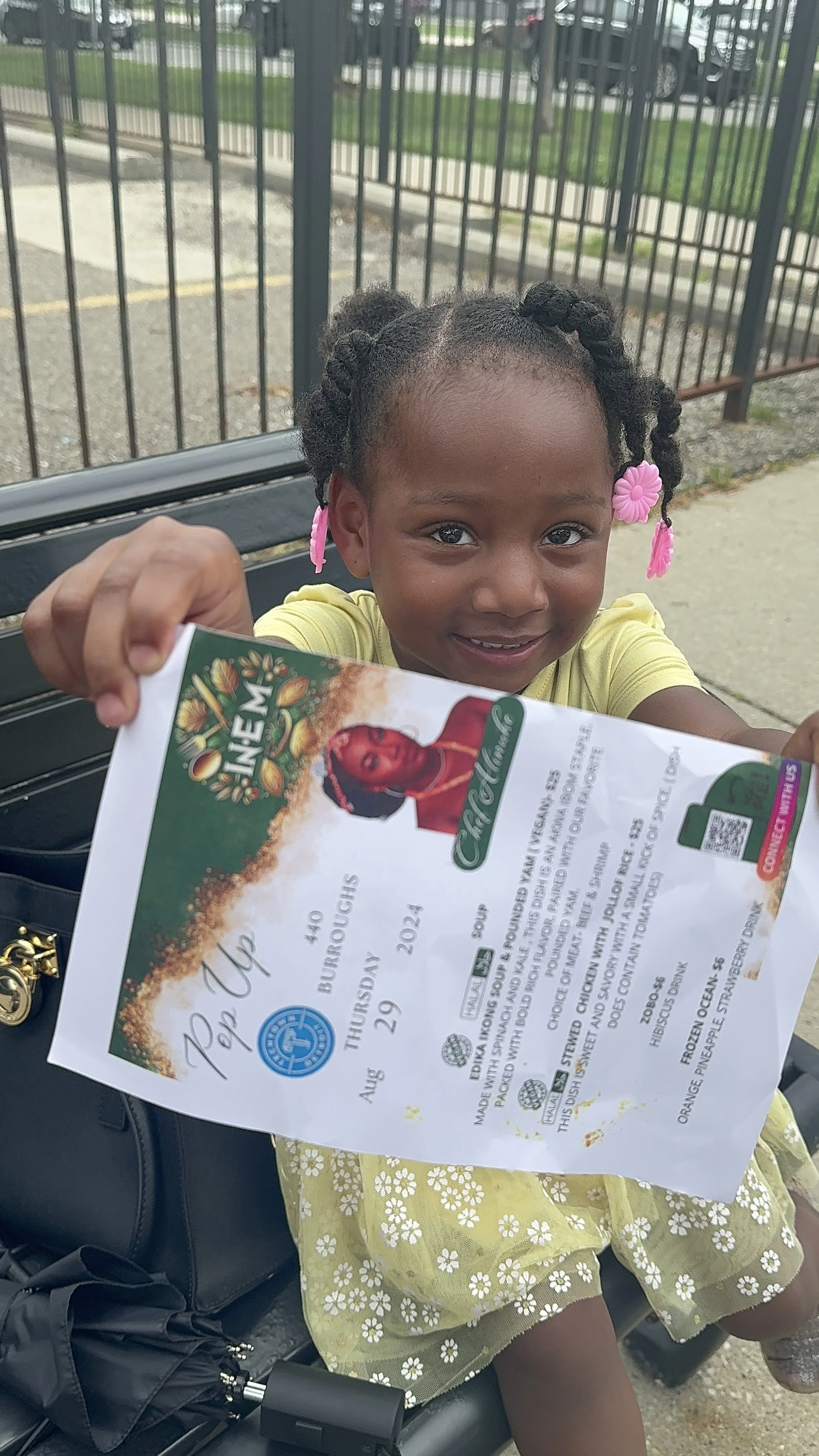 A young girl sitting on a park bench holding a ticket for an upcoming event, smiling at the camera. She has styled hair with pink and purple accessories, wearing a yellow dress with white floral patterns.