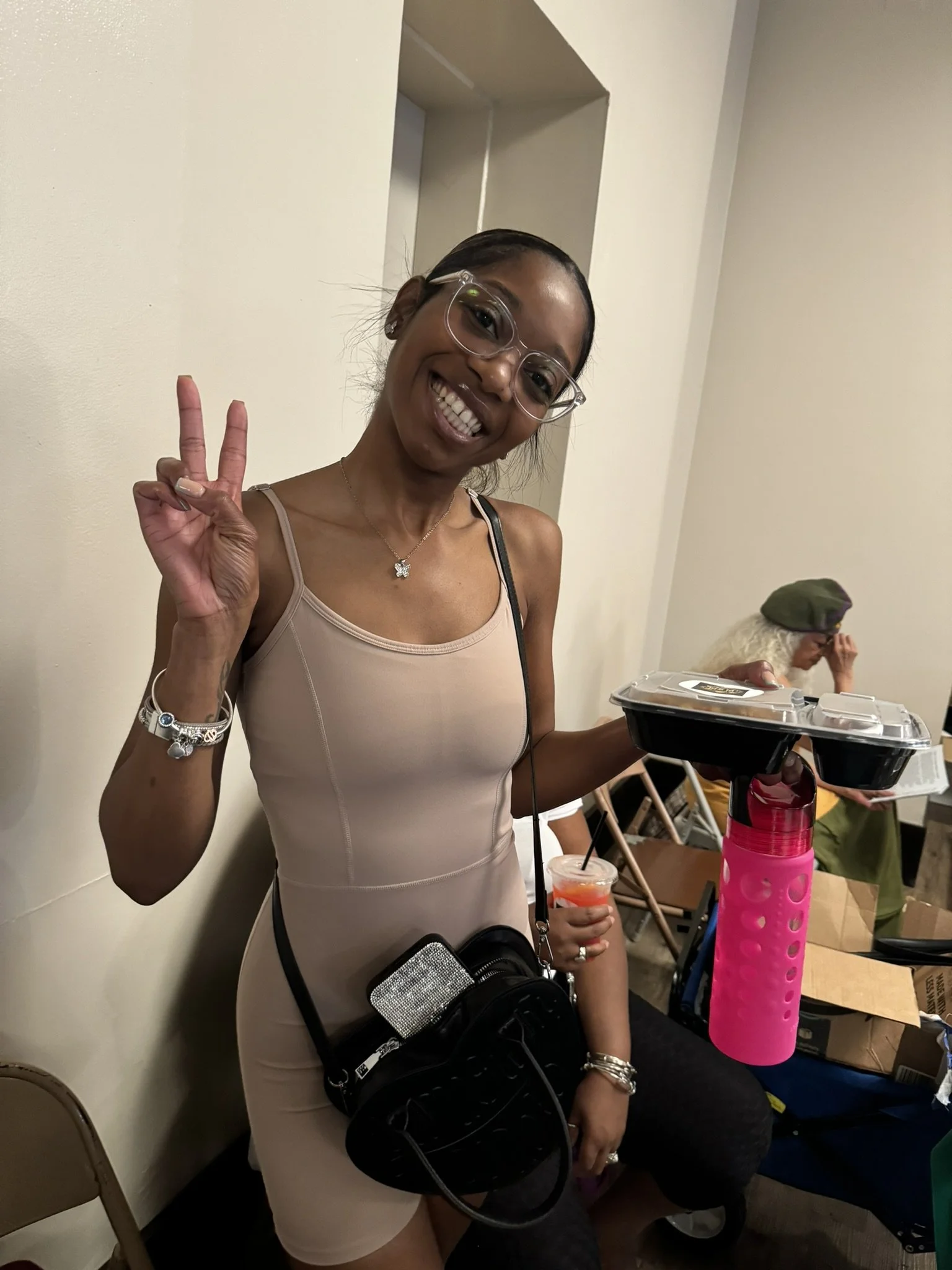 Smiling woman wearing glasses and a beige dress, making a peace sign with her right hand, holding a black food tray and a pink water bottle, with a woman sitting in the background.