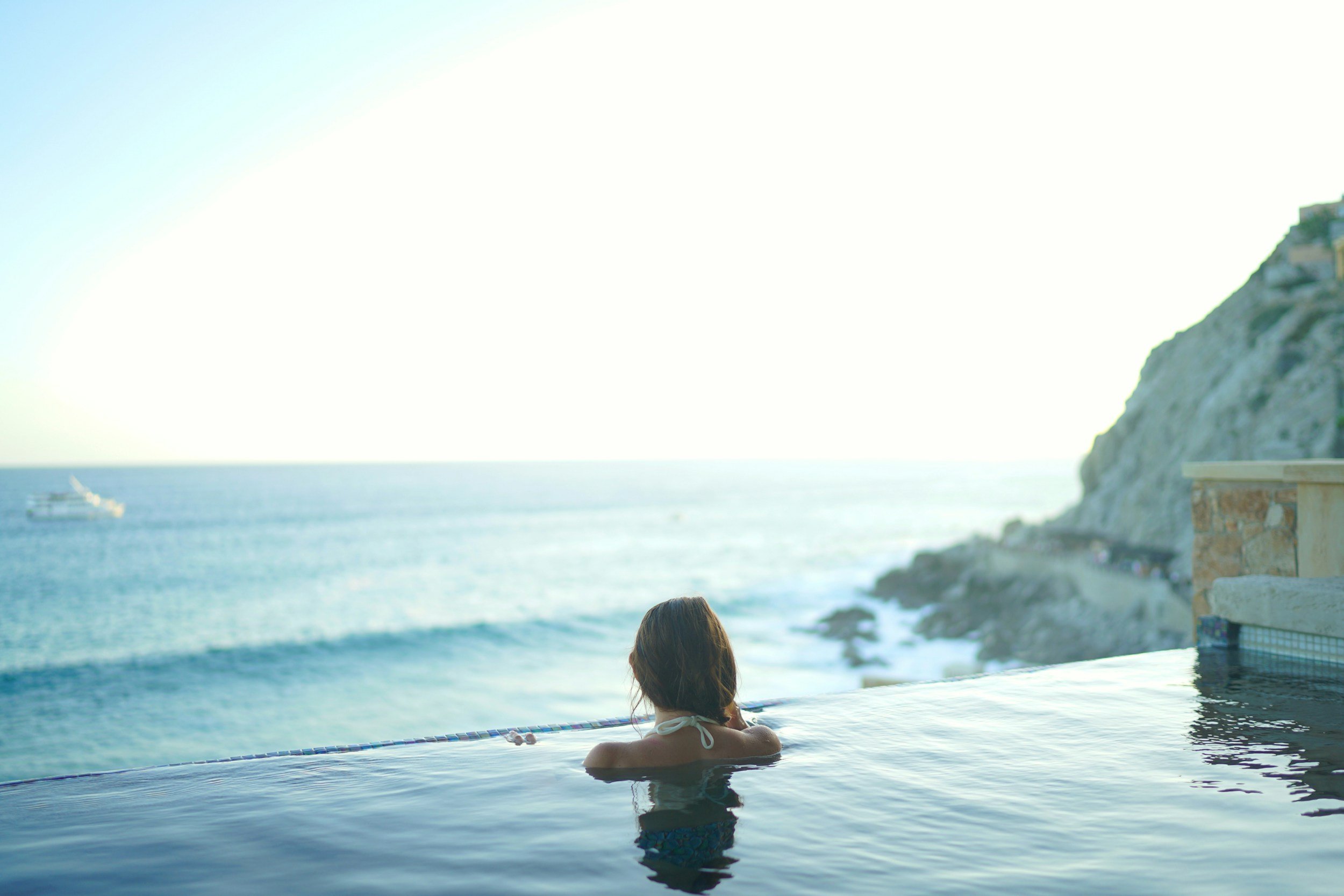 A woman relaxing in an infinity pool overlooking the ocean with cliffs and a boat in the distance during sunset.