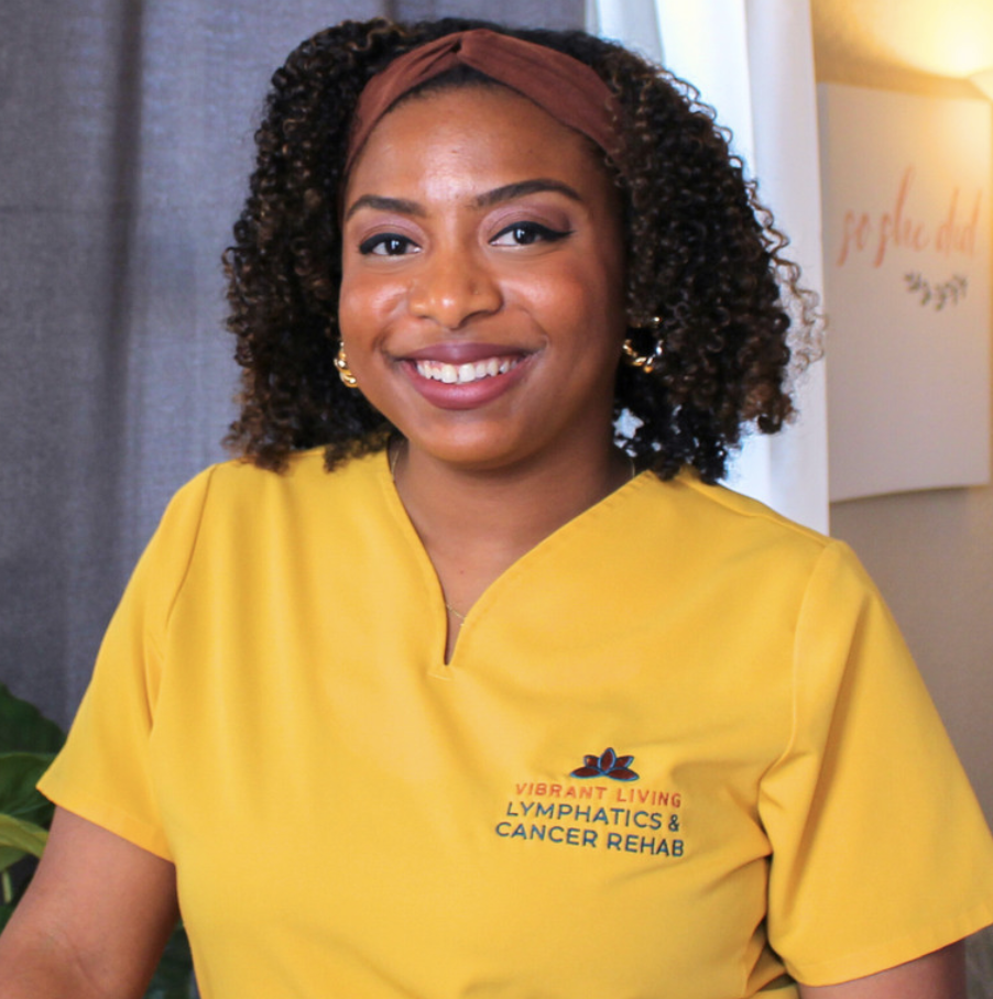 A smiling woman with curly hair wearing a yellow uniform with the text Vibrant Living Lymphatics & Cancer Rehab.