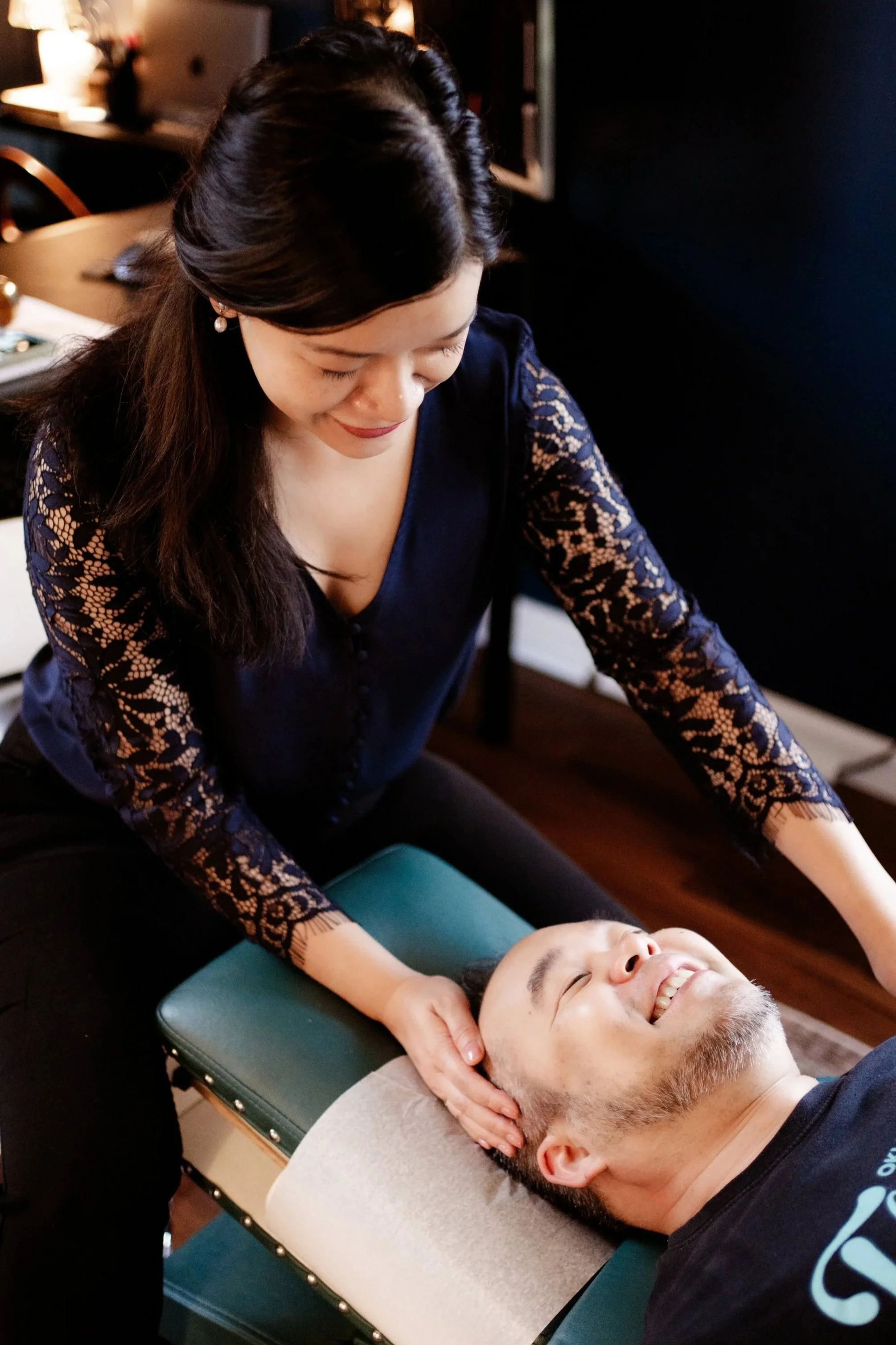 A woman is giving a neck massage to a man lying face up on a massage table, both smiling. The woman is wearing a dark blue lace top and has dark hair, while the man has a beard and is dressed in a black t-shirt.
