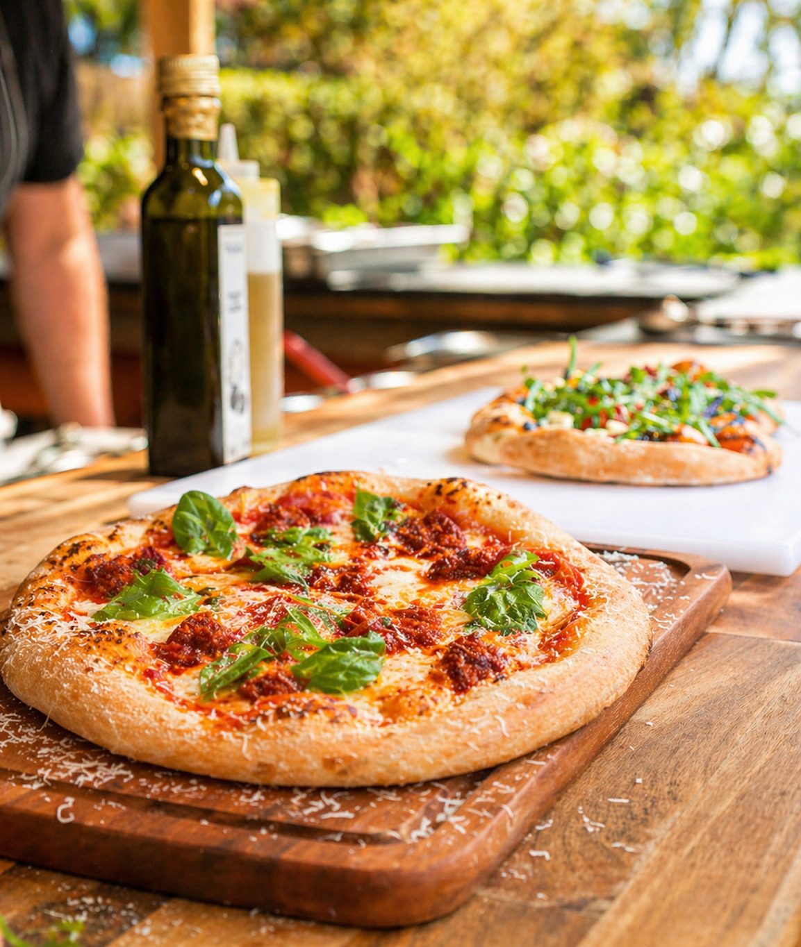 Two freshly baked pizzas with leafy greens on a wooden table, with bottles of oil and condiments in the background, outdoors setting with trees and sunlight.