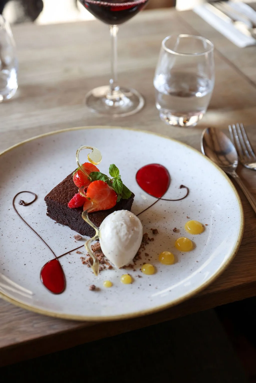 A chocolate dessert with strawberries, a scoop of vanilla ice cream, and decorative sauces on a white plate, set on a wooden table with wine and water glasses in the background.