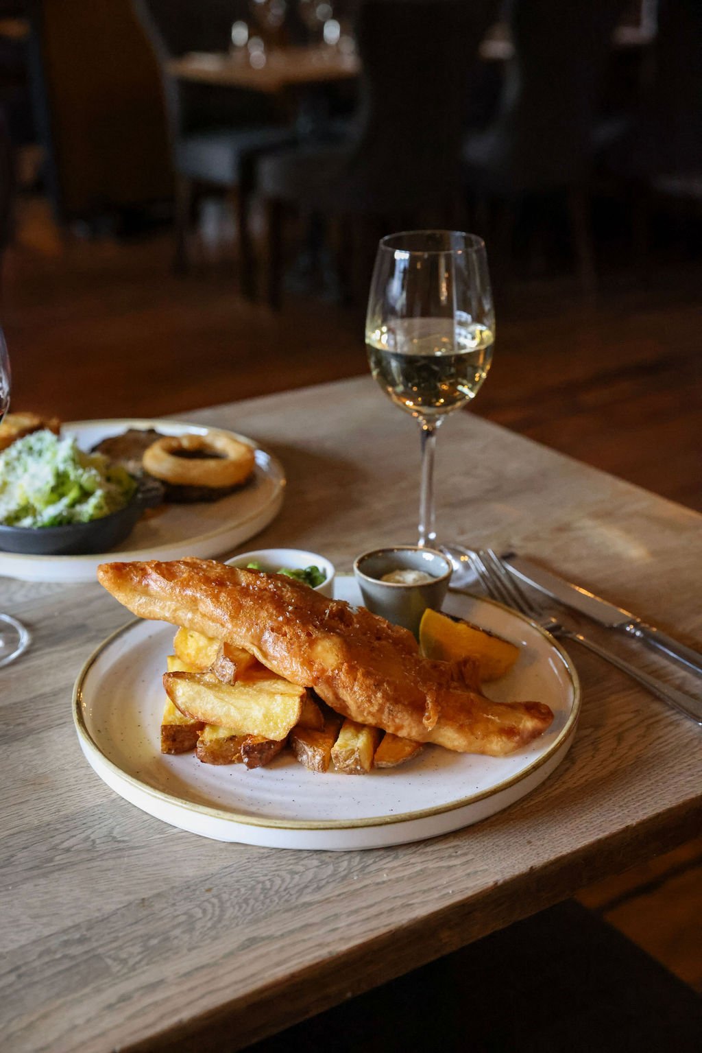 Plate with fish and chips, lemon wedges, and small bowls of tartar sauce and malt vinegar on a wooden table. Also a glass of white wine and a separate dish of salad and bread in the background.