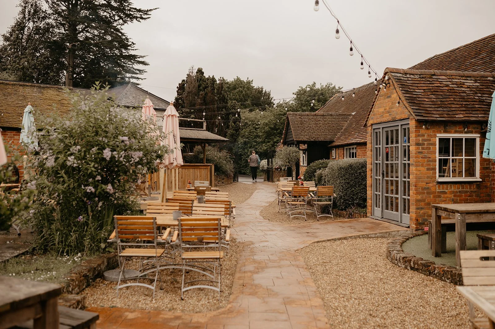 An outdoor seating area with wooden tables and chairs, surrounded by plants and shrubs, with a pathway leading to a person walking away in the distance, and a small brick building with a glass door on the right. String lights hang overhead, creating a cozy atmosphere.