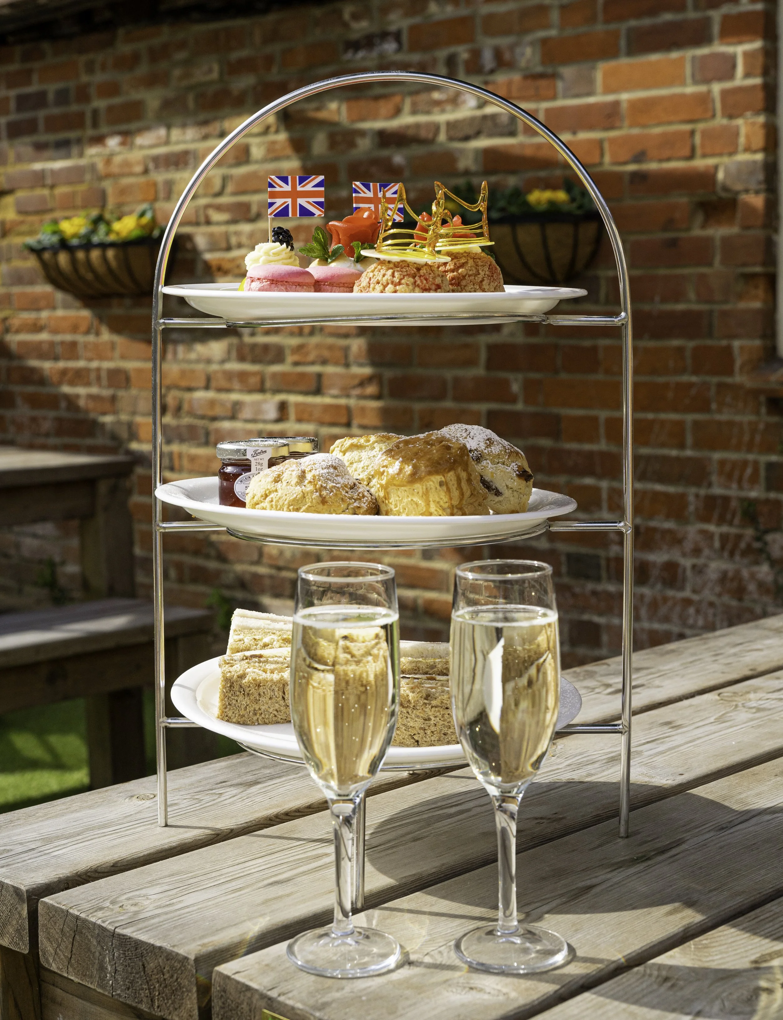 A three-tiered dessert stand with assorted desserts and two glasses of champagne on a wooden table outdoors against a brick wall. The top tier has decorated treats with Union Jack flags, the middle tier features sliced cake with powdered sugar, and the bottom tier holds pastries.