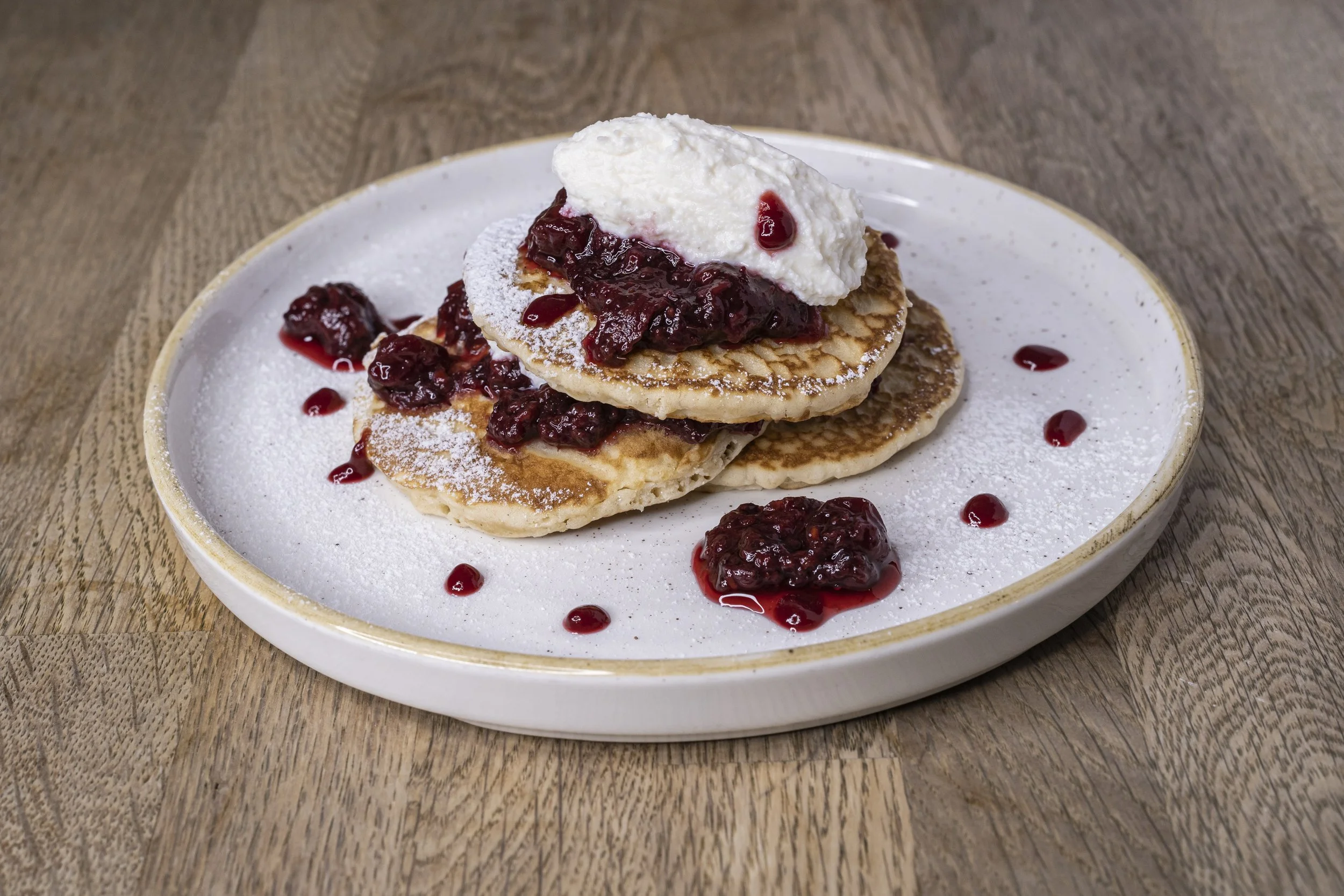 A plate of pancakes topped with cherry compote and whipped cream, with some cherry topping drizzled around on the plate.