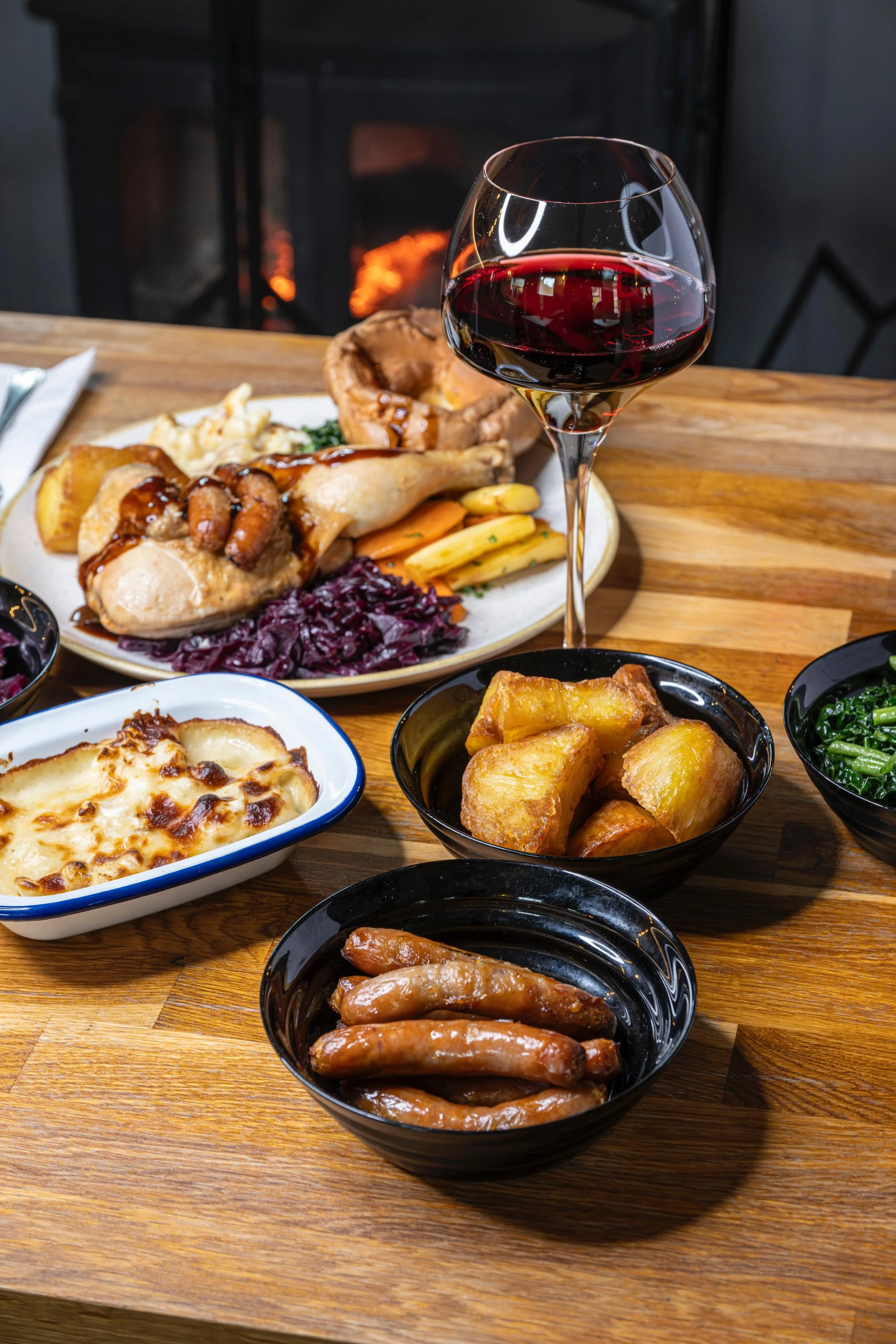 A table with various dishes including roasted chicken with gravy, fried potatoes, macaroni and cheese, a baked dish with cheese, cooked sausages, and bowls of greens. A glass of red wine is in the foreground. In the background, there's a fireplace with a fire burning.