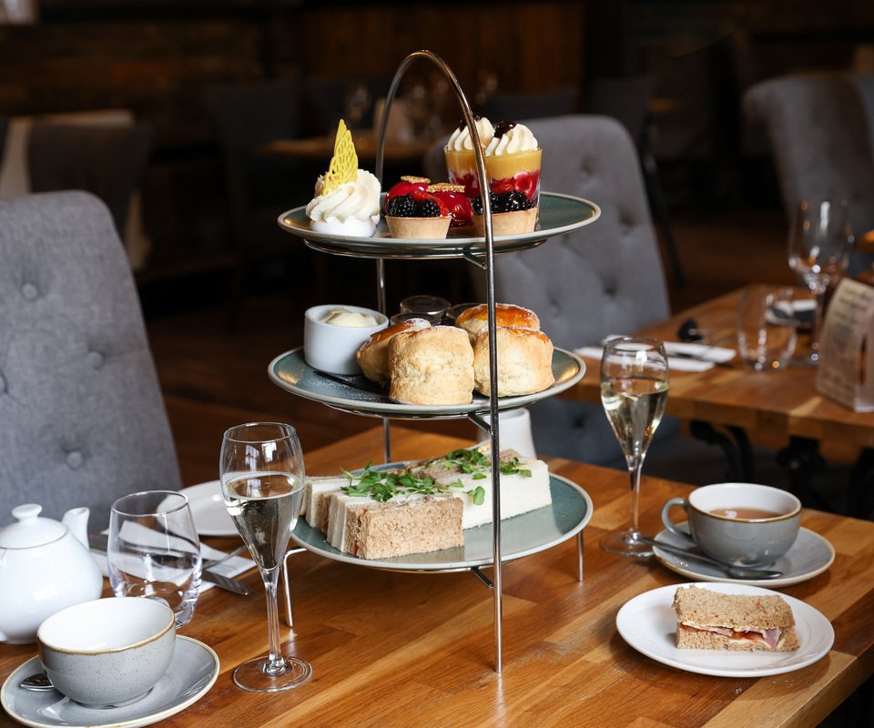 Three-tiered serving tray with assorted pastries and desserts, including scones, tarts, cakes, and cream-filled confections, set on a wooden table with tea cups, wine glasses, and a teapot in a cozy restaurant or café environment.