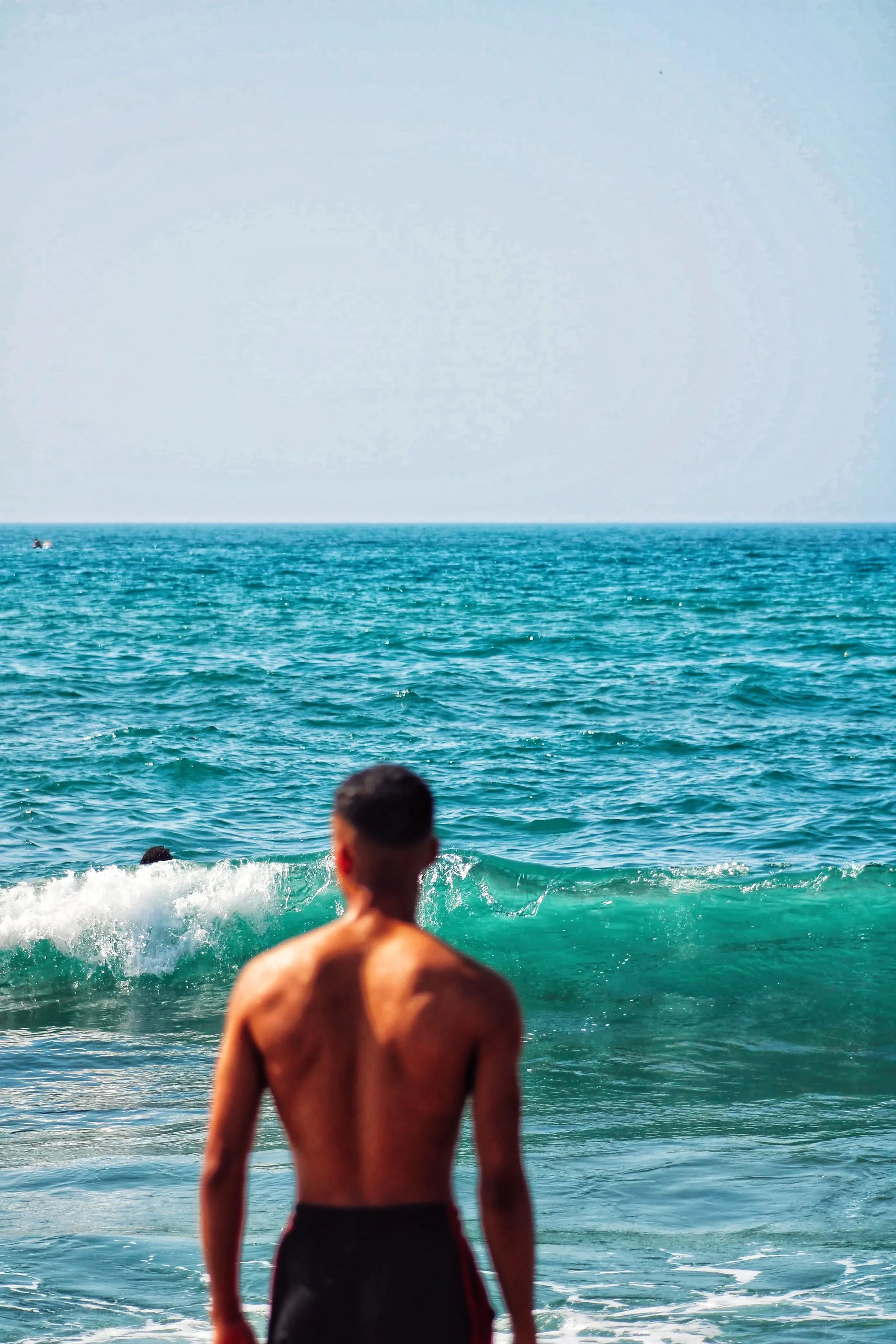 IMG1 photo of a shirtless man in a bathing suit with a bright blue body of water in focus in front of him