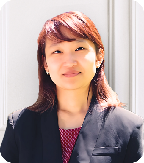 A woman with short reddish-brown hair, wearing a black blazer and a maroon patterned top, standing outdoors in front of a white door.