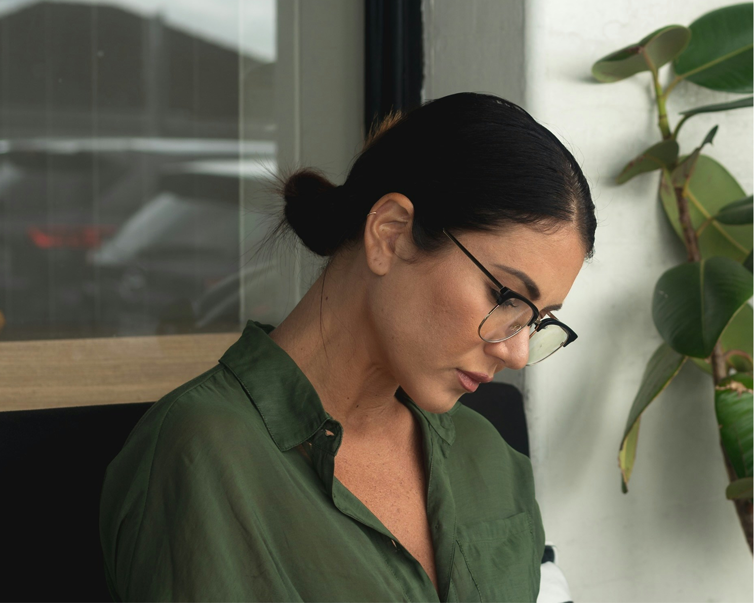 A woman with black hair tied back, wearing glasses and a green shirt, sitting indoors near a window and a potted plant.