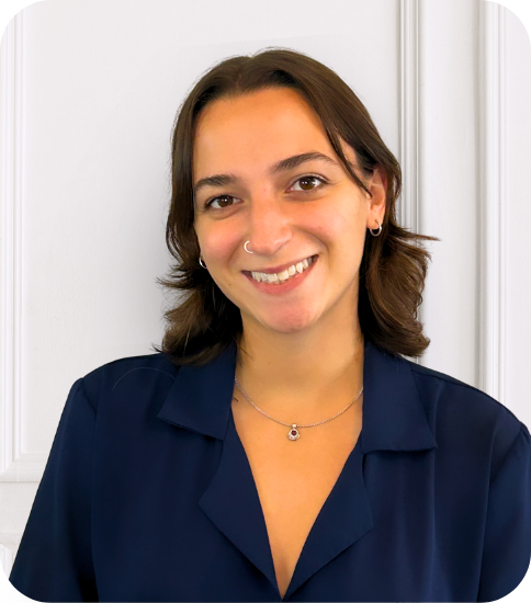 A young woman with shoulder-length brown hair, wearing a navy blue shirt, smiling, standing in front of a white wall.