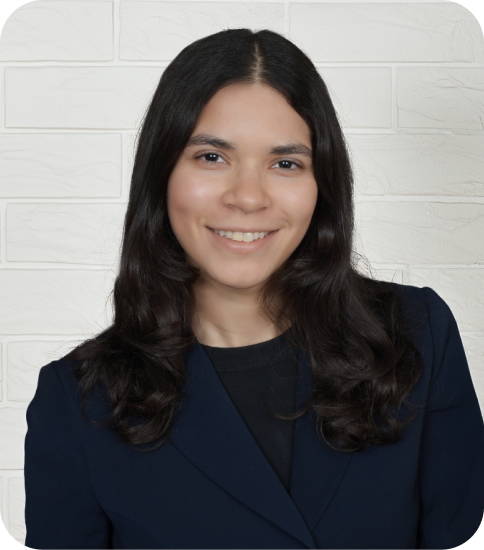 A young woman with long dark hair, smiling, wearing a navy blazer and black top, standing against a white brick wall.
