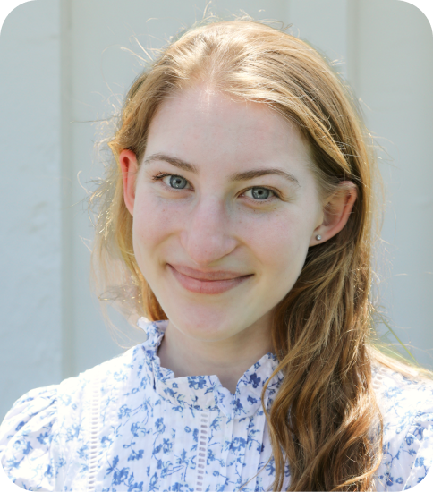 A young woman with long, light brown hair and blue eyes smiling outdoors in front of a light-colored wall, wearing a white blouse with blue floral patterns.