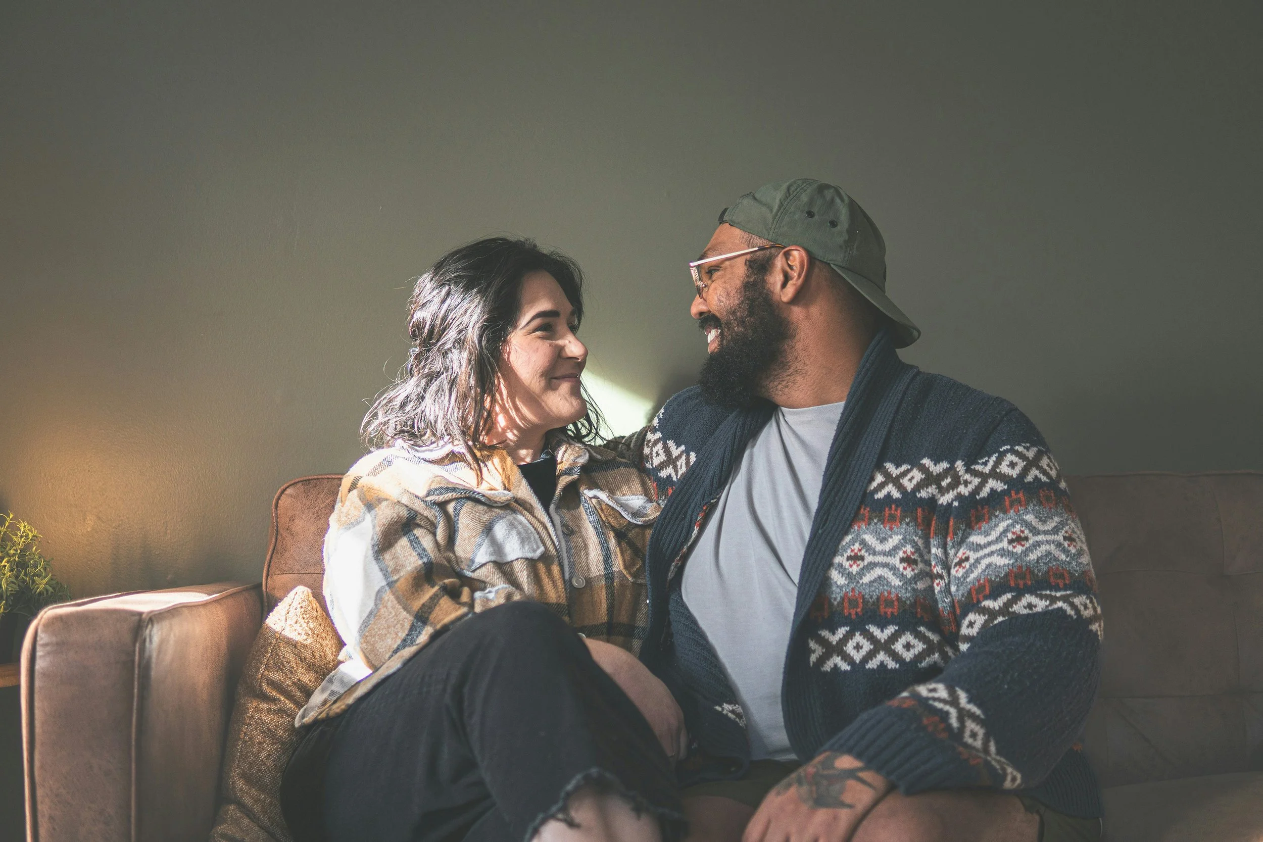 A couple sitting on a couch, smiling and looking at each other, in a cozy living room setting.